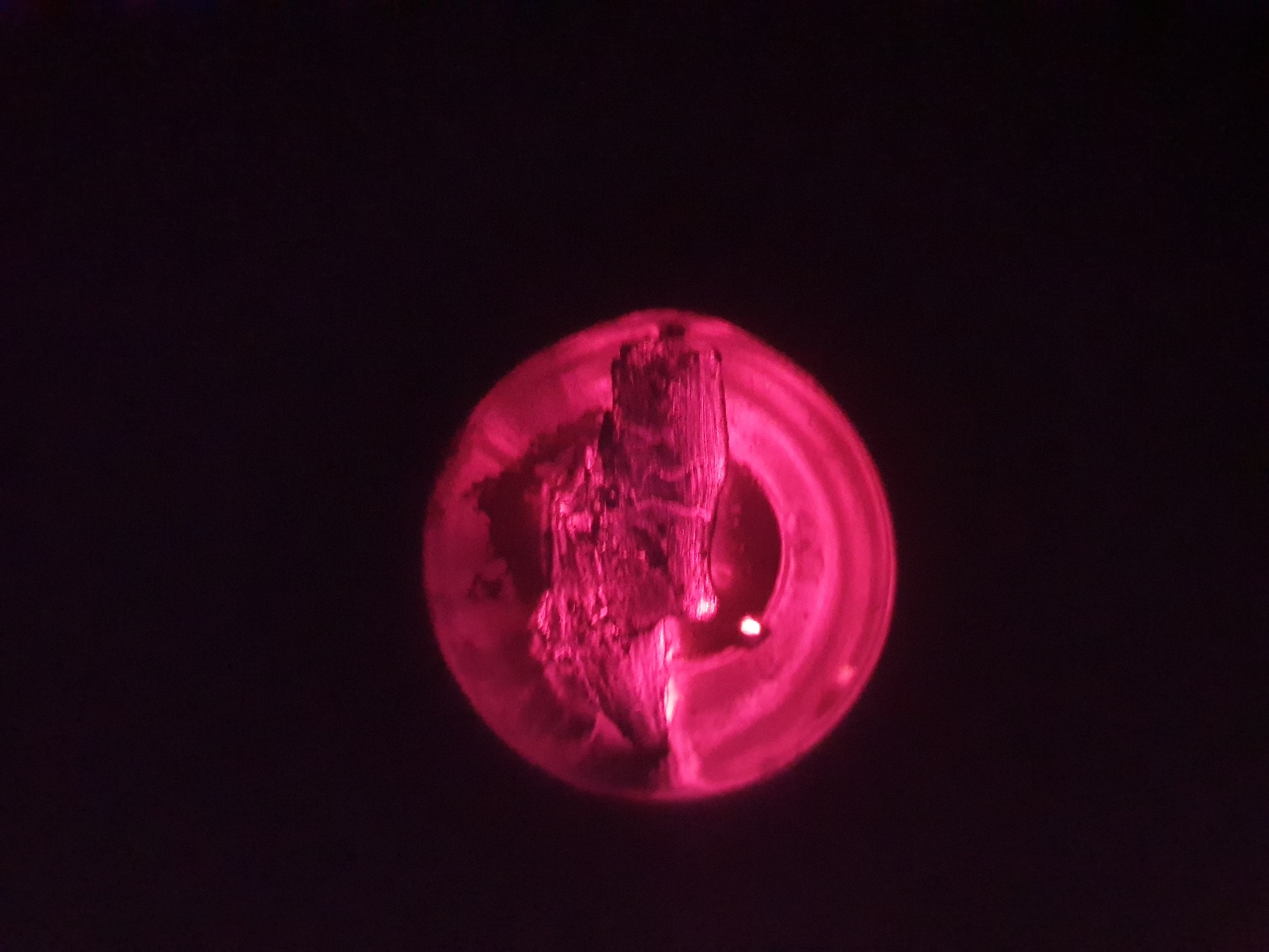 An abstract, close-up, overhead shot shows a piece of wood that has been scorched or burnt, illuminated by a vibrant pink light source, likely a bulb. The wood is positioned within a circular, possibly glass or plastic, container. The background is entirely black, creating a stark contrast with the illuminated subject. The texture of the wood is rough and splintered, indicating it has undergone significant heat exposure. The pink light casts strong shadows and highlights, emphasizing the irregular surface of the wood. The image is taken at an unknown location in an unknown city.