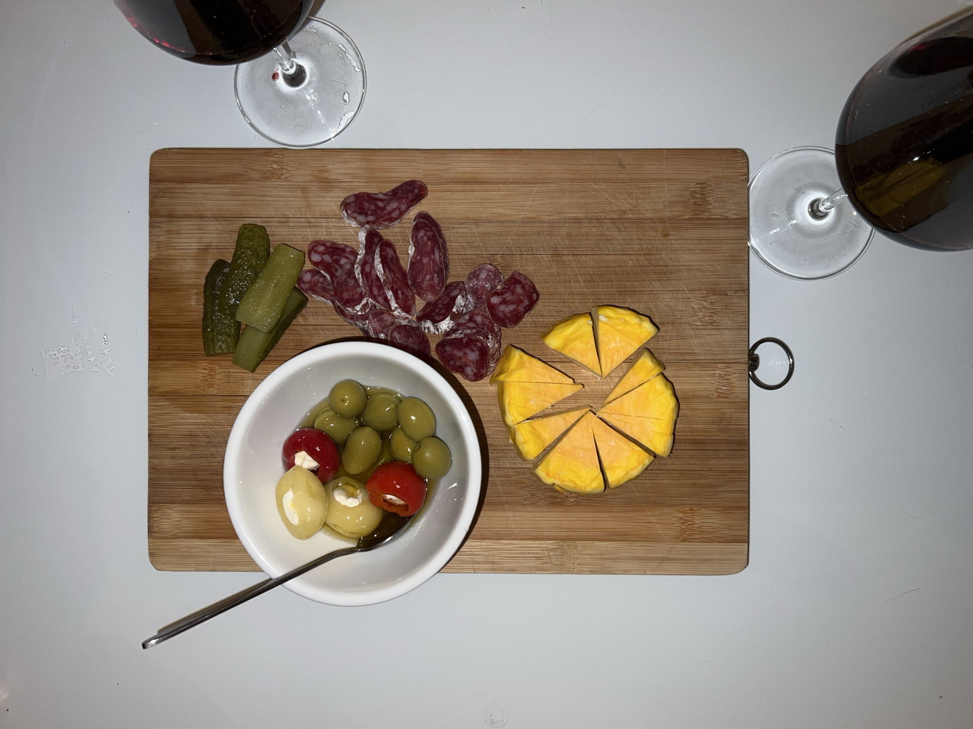 The image shows a wooden cutting board with various food items. There are sliced pickles, cut salami, a bowl of olives and stuffed peppers with a spoon, and a wheel of yellow cheese cut into slices. Two glasses of red wine are visible on the sides of the board. The background is a plain white surface. The location in the city of Bar, Montenegro is not identifiable from the information in the image.