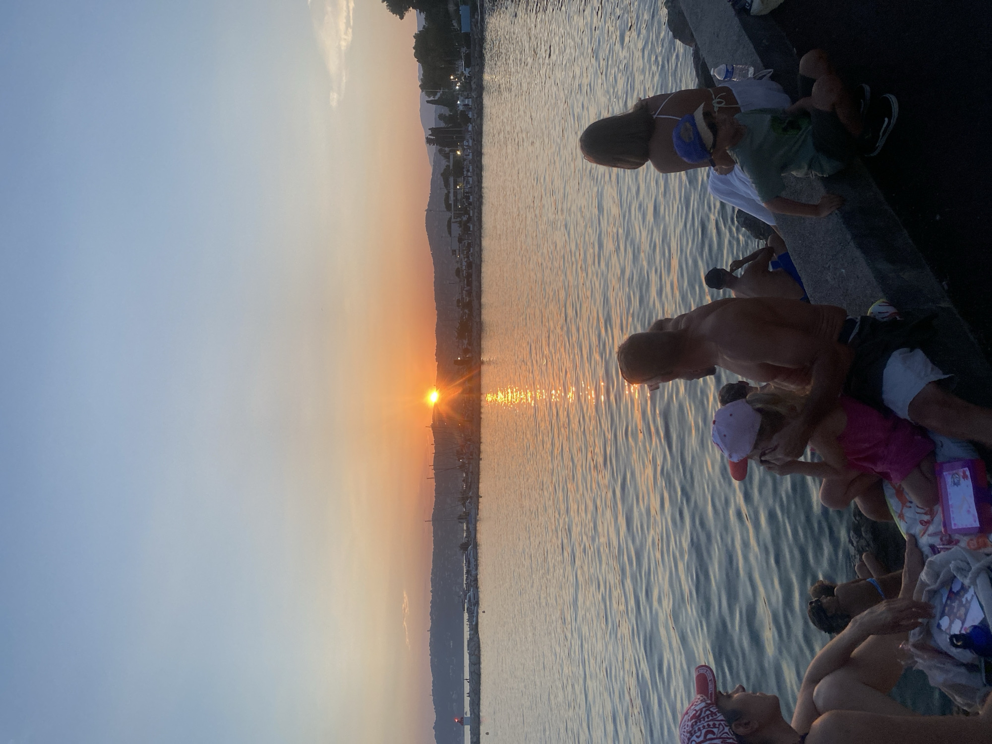 The image depicts a group of people gathered by the water at sunset in Antibes, France. The sun is low on the horizon, casting a warm, golden glow across the water and the sky. The scene is set along a rocky embankment overlooking the sea. Several individuals, including adults and children, are seated or lounging on the embankment. One person is wearing a blue baseball cap, and another is wearing a pink and white baseball cap. There are various objects scattered around, including towels and bags. The water appears calm with gentle ripples reflecting the sunset. In the background, a coastline with buildings and hills is visible, with a few boats docked in a harbor. The overall atmosphere suggests a relaxed and peaceful evening gathering, enjoying the scenic beauty of the sunset. There are no visible text elements in the image.