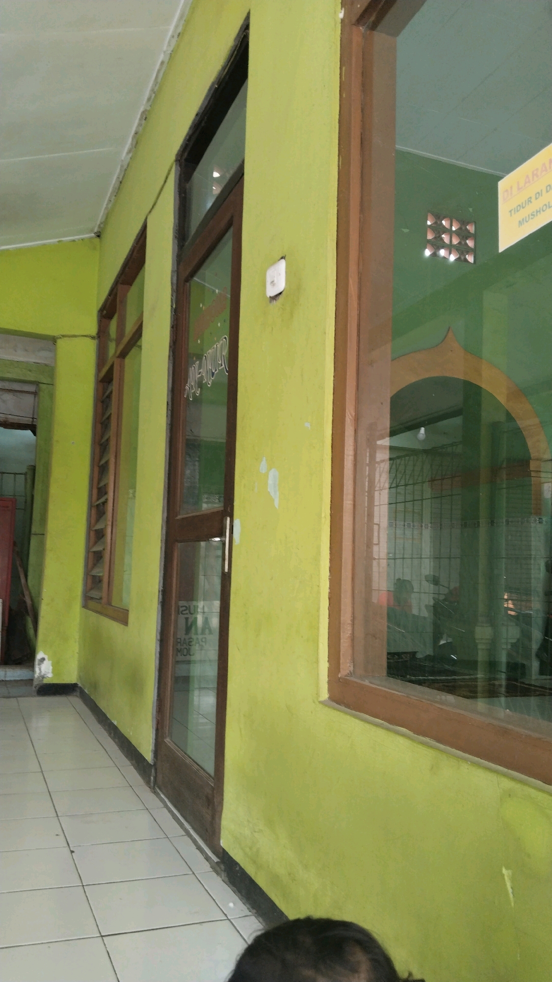 The image shows a hallway with lime green walls. On the right side of the image, there is a large window with a brown wooden frame. The window looks into a room with green walls and an arched doorway. A sign with yellow lettering is visible through the window. On the left side of the image, there is a closed wooden door with a glass panel. The floor is tiled with white square tiles. The ceiling is white. The overall impression is of an interior hallway or corridor.