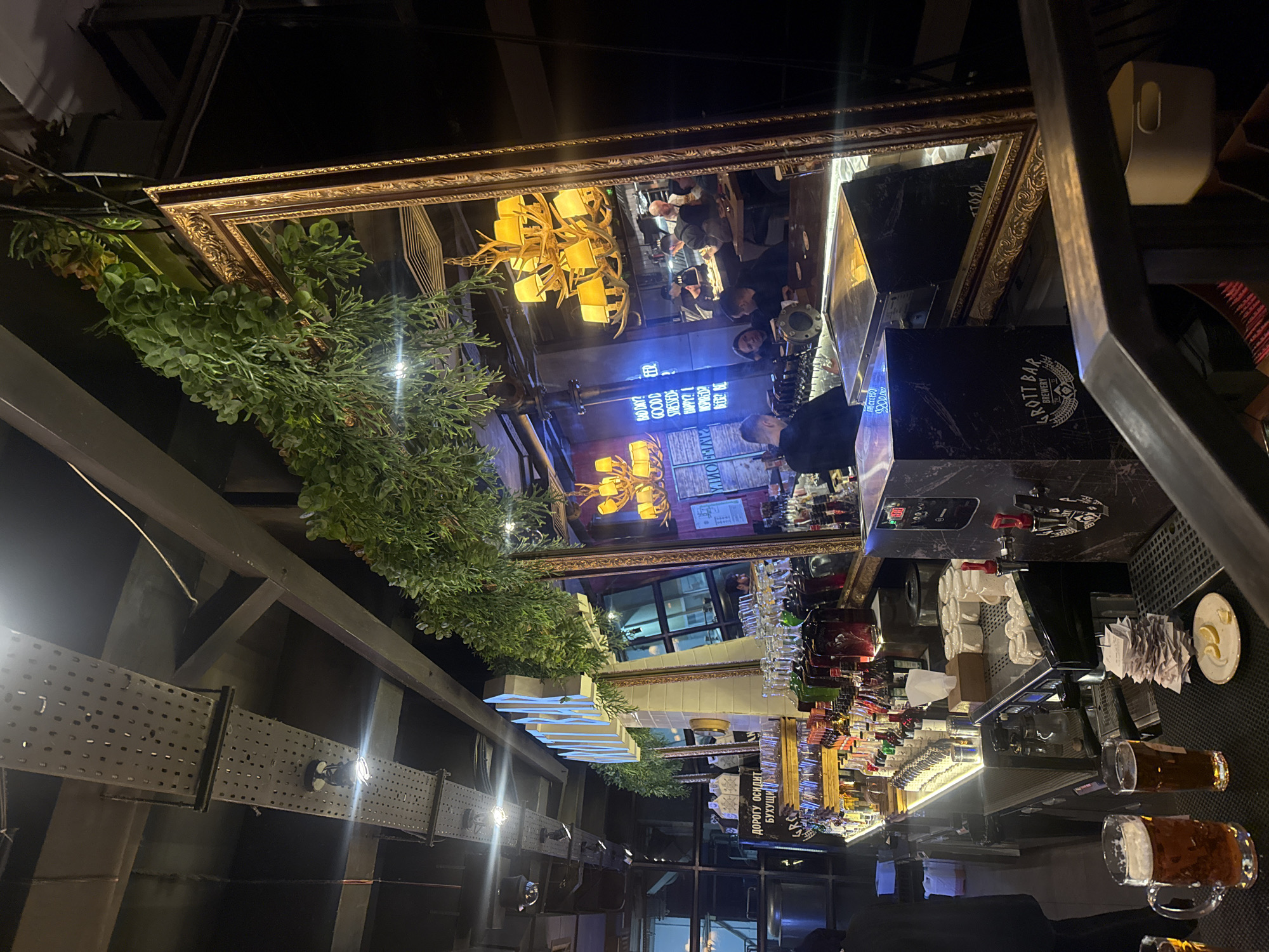 The image captures an indoor nighttime scene within a modern bar or pub, located in Yekaterinburg, Russia. The setting features an industrial-chic aesthetic with exposed metal structures and dark walls, enhanced by artificial lighting.
A dominant feature is a large, ornate golden-framed mirror installed on the ceiling, reflecting the lively space below. Visible in this reflection are several patrons, some seated at tables or the bar, and a person appearing to be a bartender. One individual in the reflection wears a dark top and a mask.
The bar area is well-stocked with numerous bottles of various spirits, rows of inverted glasses, and a modern beer tap system. Several mugs filled with amber-colored beer are visible on the dark bar counter, alongside a small plate with lemon wedges. Other equipment, possibly for coffee, is also present behind the bar.
Decorative elements include artificial green plants, resembling eucalyptus and cypress, draped along the edges of the ceiling mirror. Industrial-style track lighting is mounted on perforated metal panels on the walls and ceiling structures. A distinctive chandelier, designed with antler-like shapes and yellow lampshades, is prominently reflected in the mirror.
The overall activity suggests patrons socializing and enjoying drinks in a relaxed atmosphere.
Visible text includes:
- Reflected in the ceiling mirror, a neon sign displays phrases such as "GOOD BEER," "STRESSED? HAPPY? WORKED? DRINK BEER." Other reflective text appears to read "RIVER OIL" and the numerical "100000."
- On a black surface at the bar, the text "GROTT BAR BREWERY" is seen, accompanied by a logo.
- A white sign further down the bar area shows Russian text: "ДОРОГУ ОСИДИТ БУХУЩИ."