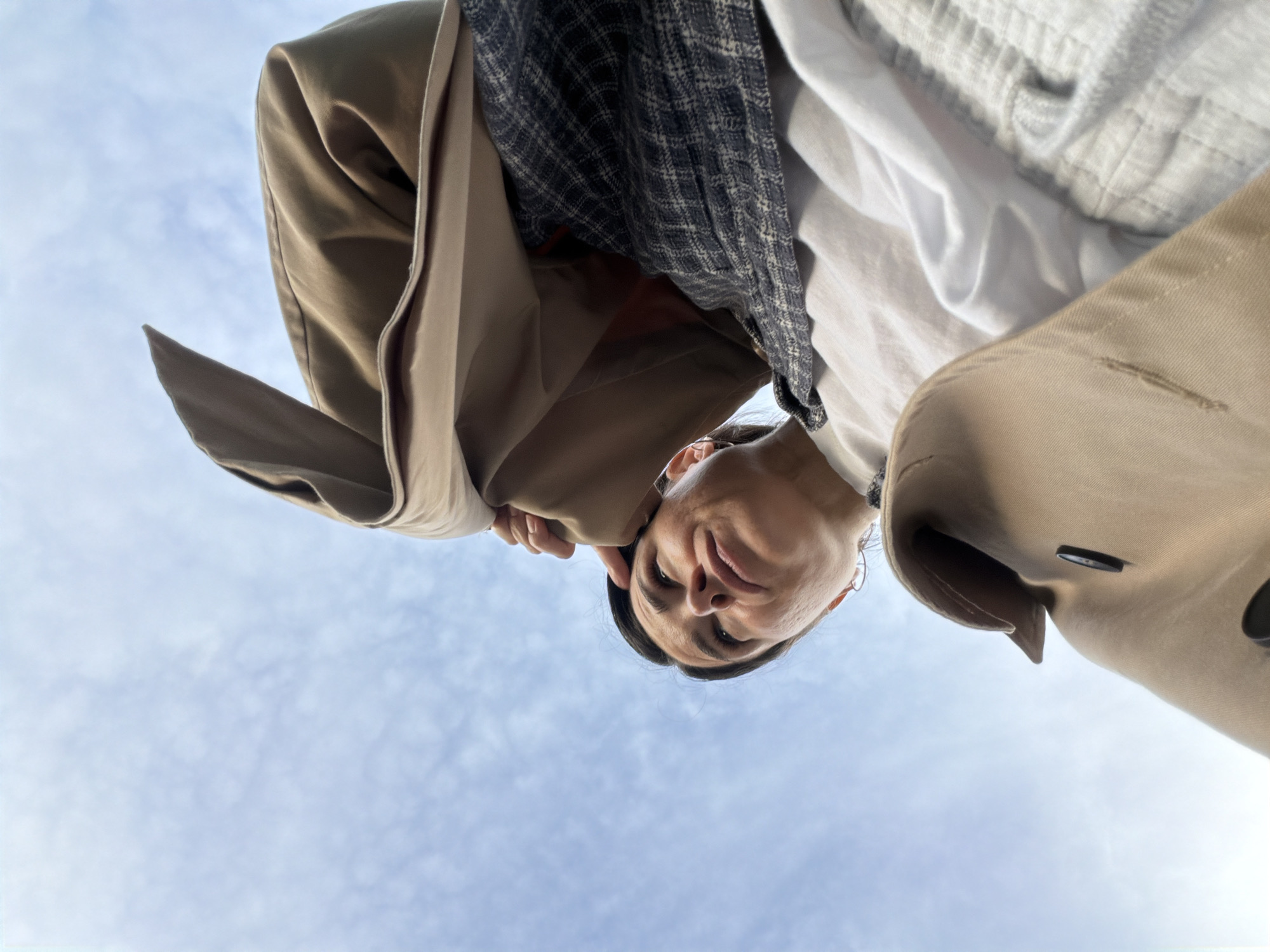 The image, taken from a low-angle perspective against a backdrop of a light blue, partly cloudy sky, features a person looking down at the camera. The individual is wearing a tan trench coat, a white collared shirt, and a black and grey checkered garment underneath. The person's face is visible, with dark hair pulled back. The lighting suggests an outdoor environment. It is not possible to confirm the specific location within Budva, Montenegro based on the visual data alone.