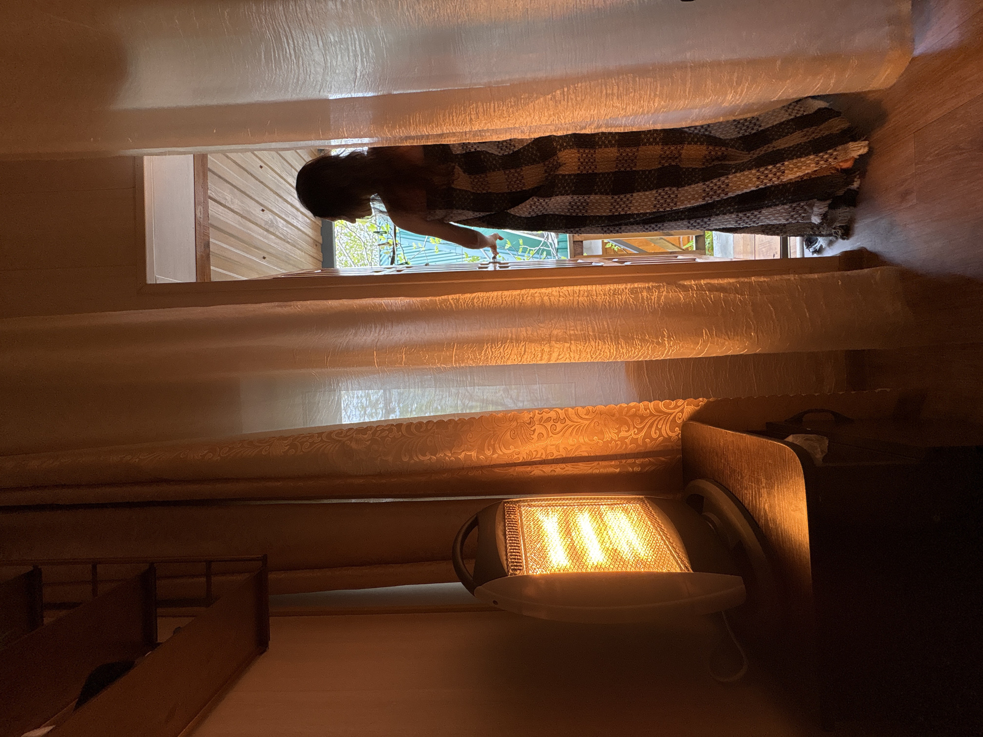 A person wrapped in a checkered blanket is seen opening a window. The scene is indoors, with the lower half of the image dominated by a space heater. A staircase can be seen in the lower left. The location appears to be Gelendzhik, Russia, based on circumstantial visual clues: the architecture and the style of the window and blanket suggest a possible geographical location.