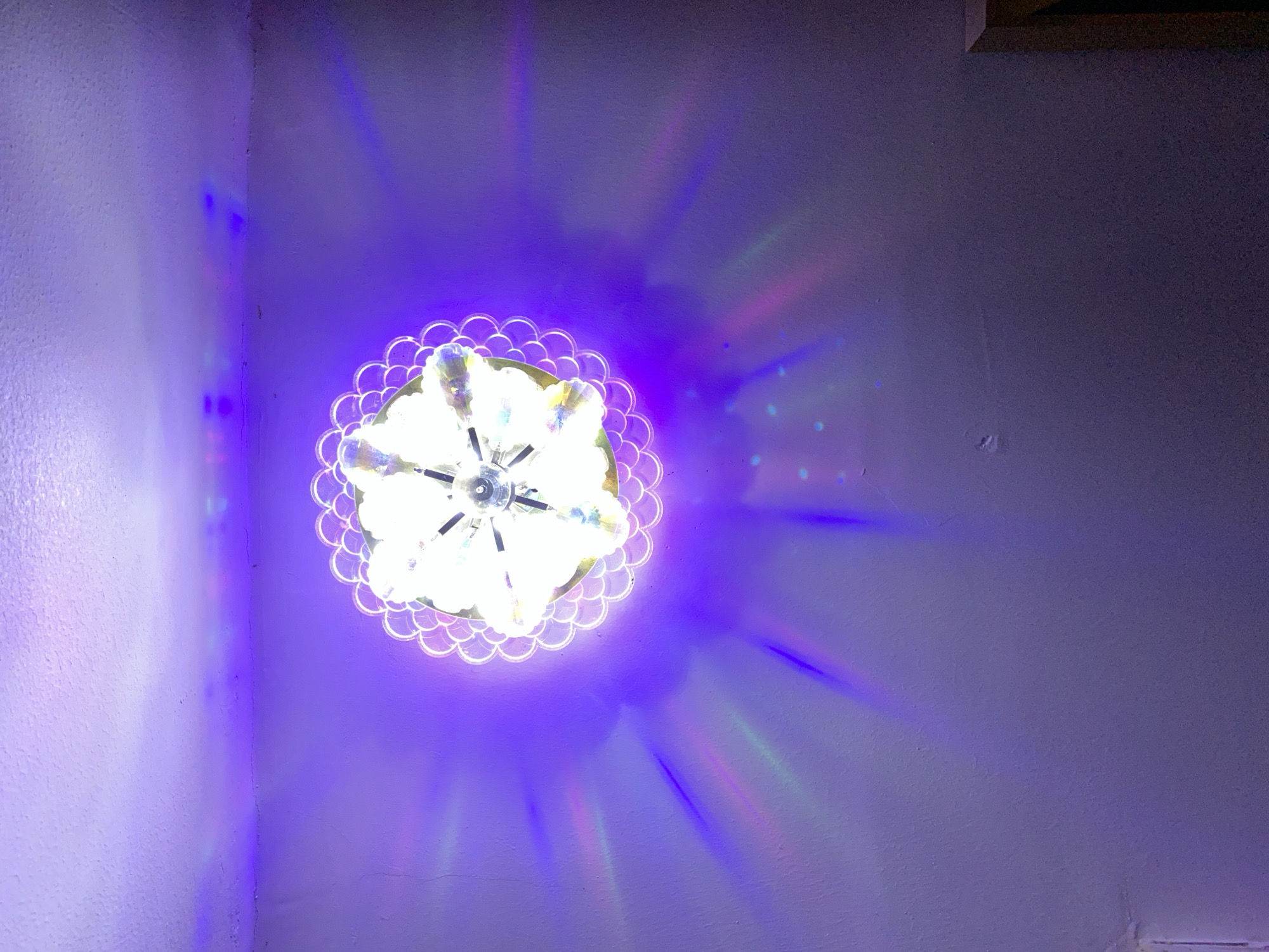 The image shows a ceiling light fixture with a decorative, multi-faceted design that projects colored light onto the surrounding wall and ceiling. The light fixture appears to be illuminated by multiple bulbs, creating a disco-ball-like effect. The walls and ceiling are painted a light purple color. The light emanating from the fixture casts colorful beams and patterns, primarily in shades of purple, pink, and green, across the surfaces. The scene is indoor and appears to be a room. There are no people or discernible activities visible in the image. The location is not provided in the image, so it cannot be determined if this is in Giginyu, Nigeria or any other city.