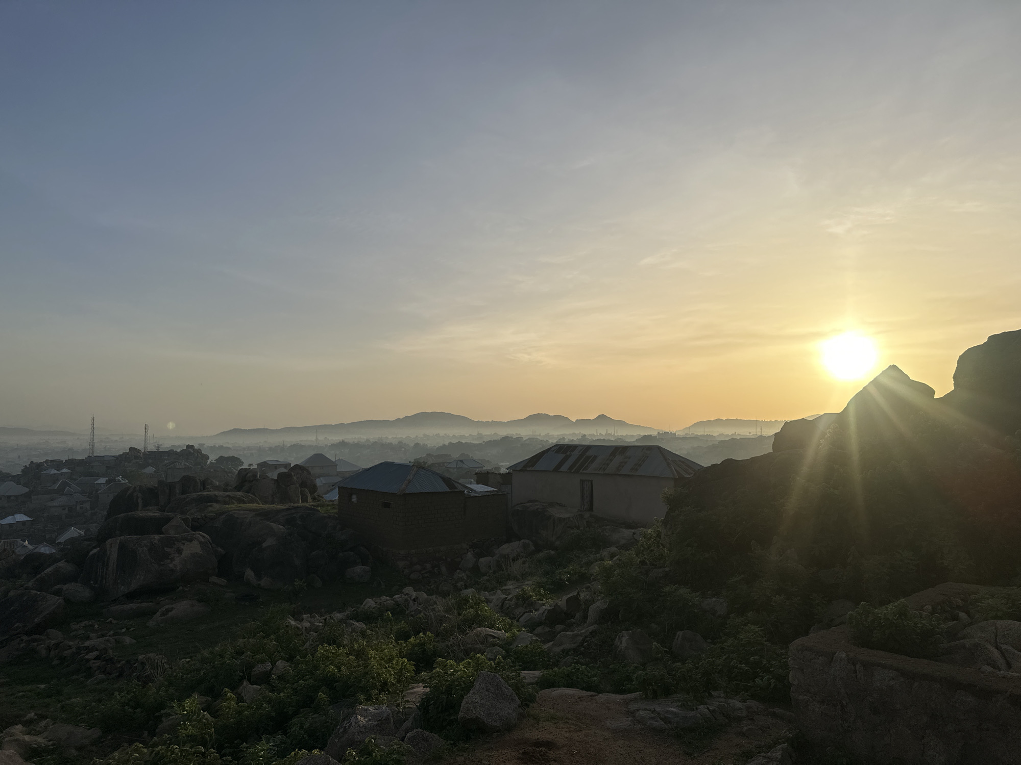 This media file captures a serene, expansive landscape bathed in the warm glow of either sunrise or sunset, specifically in Jos, Nigeria, as indicated by the location context and characteristic rock formations. The sun, a brilliant orb, is positioned on the right side of the frame, partially obscured by a dark rock outcrop, emitting strong rays that create a distinct lens flare effect.
The sky transitions from a soft, clear light blue at the top left to a rich, hazy yellow and orange towards the horizon, indicating the early morning or late evening light. Below the horizon, a widespread settlement or town stretches into the distance, partially veiled by a gentle mist or haze, with distant mountains or hills forming a faint silhouette against the bright sky.
In the midground, two single-story houses with corrugated metal roofs are prominent. One house is light-colored, while the other, to its left, is darker, appearing to be constructed from concrete blocks or mud bricks. Both are nestled among numerous large, dark, weathered rock formations that dominate the foreground and parts of the midground. These rocks are interspersed with patches of green vegetation, including bushes and small plants, adding texture to the terrain.
No people or explicit activities are visible, contributing to the tranquil atmosphere. The entire scene is characterized by dramatic backlighting, casting many foreground elements into shadow and highlighting the atmospheric haze, giving the image a sense of peace and natural beauty. There is no visible text in the image.