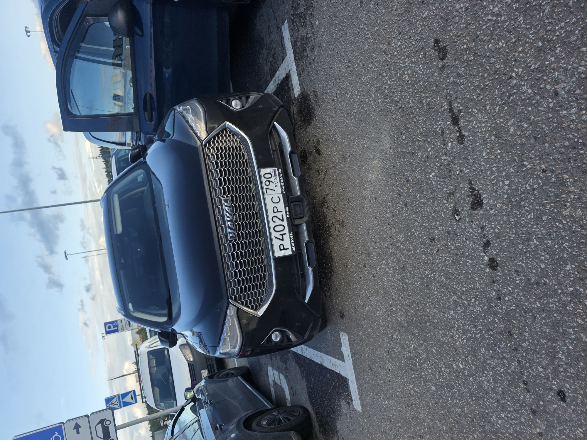This image shows a black Haval SUV parked on asphalt in Sholokhovo, Russia. The car is angled, with its front bumper and grille prominently displayed. The license plate is visible and reads "P402PC 790 RUS". Text on a sticker below the license plate indicates "+7 (495) 956-13-00" and "Москва".
Other vehicles are visible in the background, including a dark blue SUV with its door open and a white van. Several road signs are also present, including parking signs with a blue "P" symbol and arrows, and a pedestrian crossing sign. The sky is overcast with some clouds, suggesting it might be daytime but not necessarily sunny. The scene appears to be a parking lot, likely in a public area given the presence of multiple vehicles and signage. There are no people visible in the image. The asphalt shows some dark stains, possibly oil or other fluid leaks. The overall impression is a utilitarian, everyday scene.