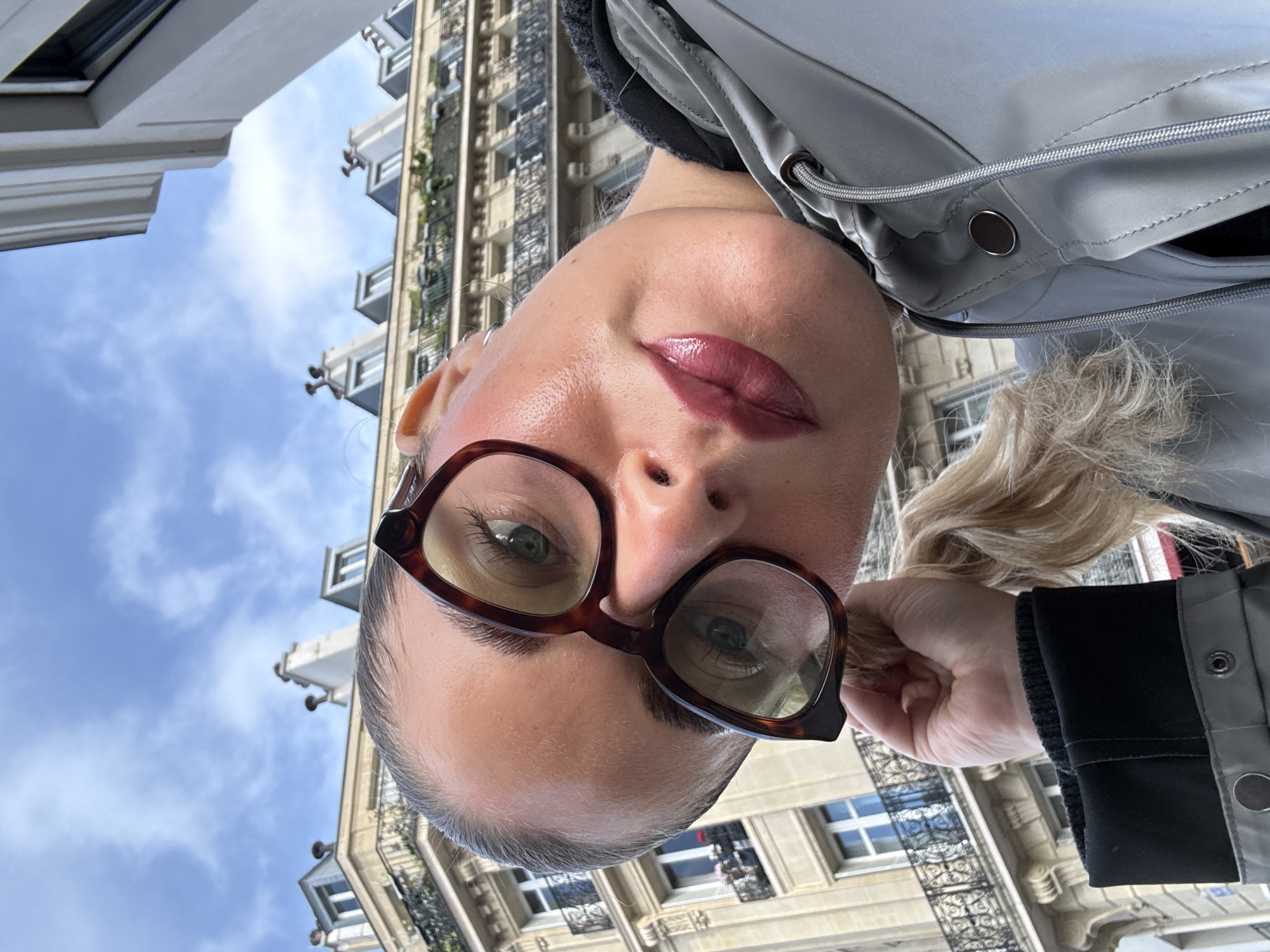 The image is a low-angle selfie of a woman wearing brown-framed glasses, with fair skin and blond hair partially pulled back. She is wearing a grey jacket with a black collar and cuffs. Her lips are a deep plum color. The background shows a street scene in Paris, France. The architecture includes ornate buildings with balconies and windows visible against a blue sky with some clouds. The buildings suggest a traditional Parisian style. The perspective indicates the photo was taken looking upwards towards the buildings.