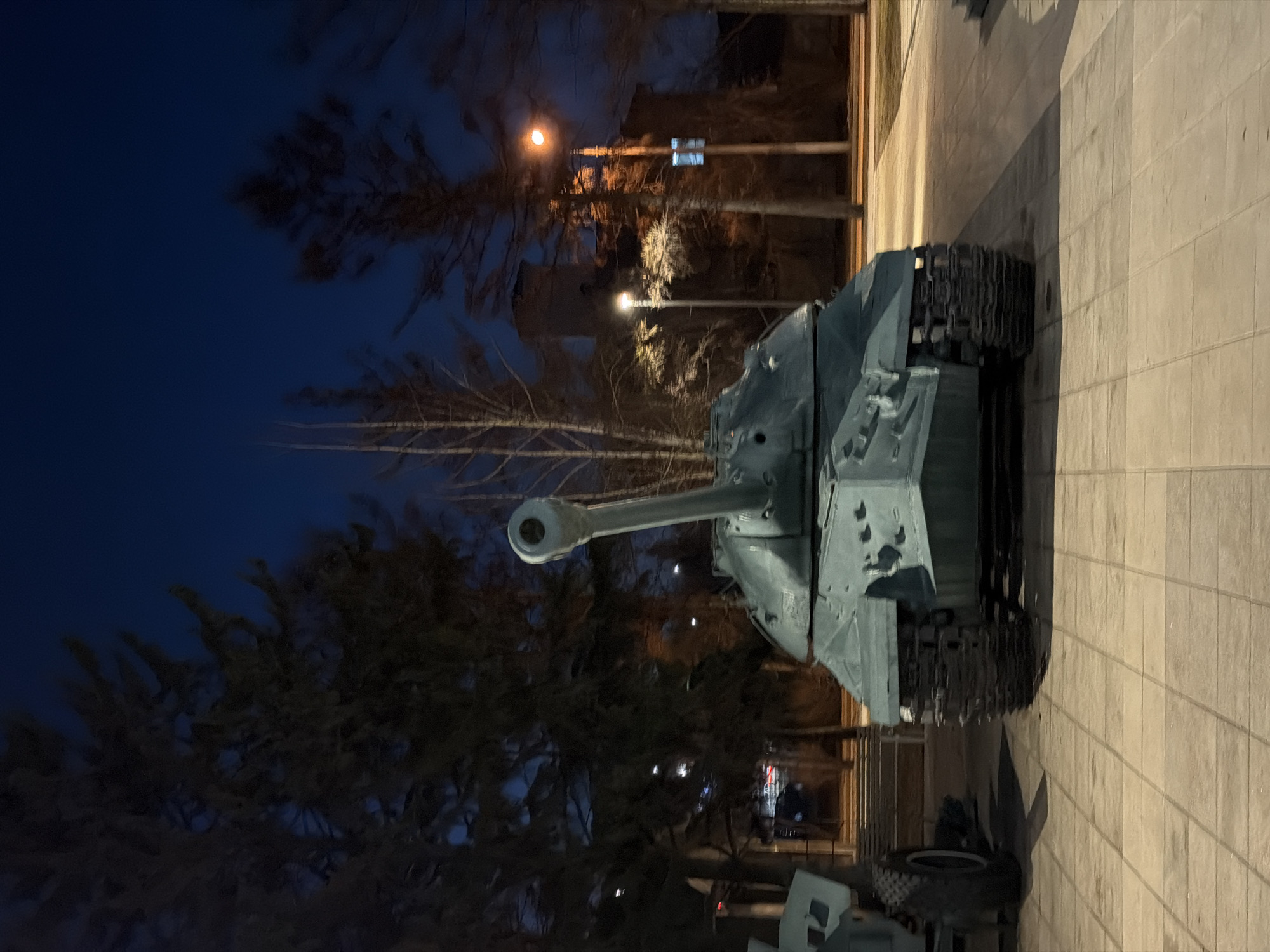 The image shows a T-34 tank, a Soviet medium tank, displayed on a paved surface in Krasnoyarsk, Russia. The tank, painted in a light gray color, is positioned with its cannon pointing towards the left side of the frame. Behind the tank are several bare trees, illuminated by streetlights. The background includes a dark blue sky, indicating it is nighttime. There are some buildings visible, but they are partially obscured by the trees. A concrete wall runs along the right side of the image. Another military vehicle, also in gray, is situated behind and partially beneath the T-34.
