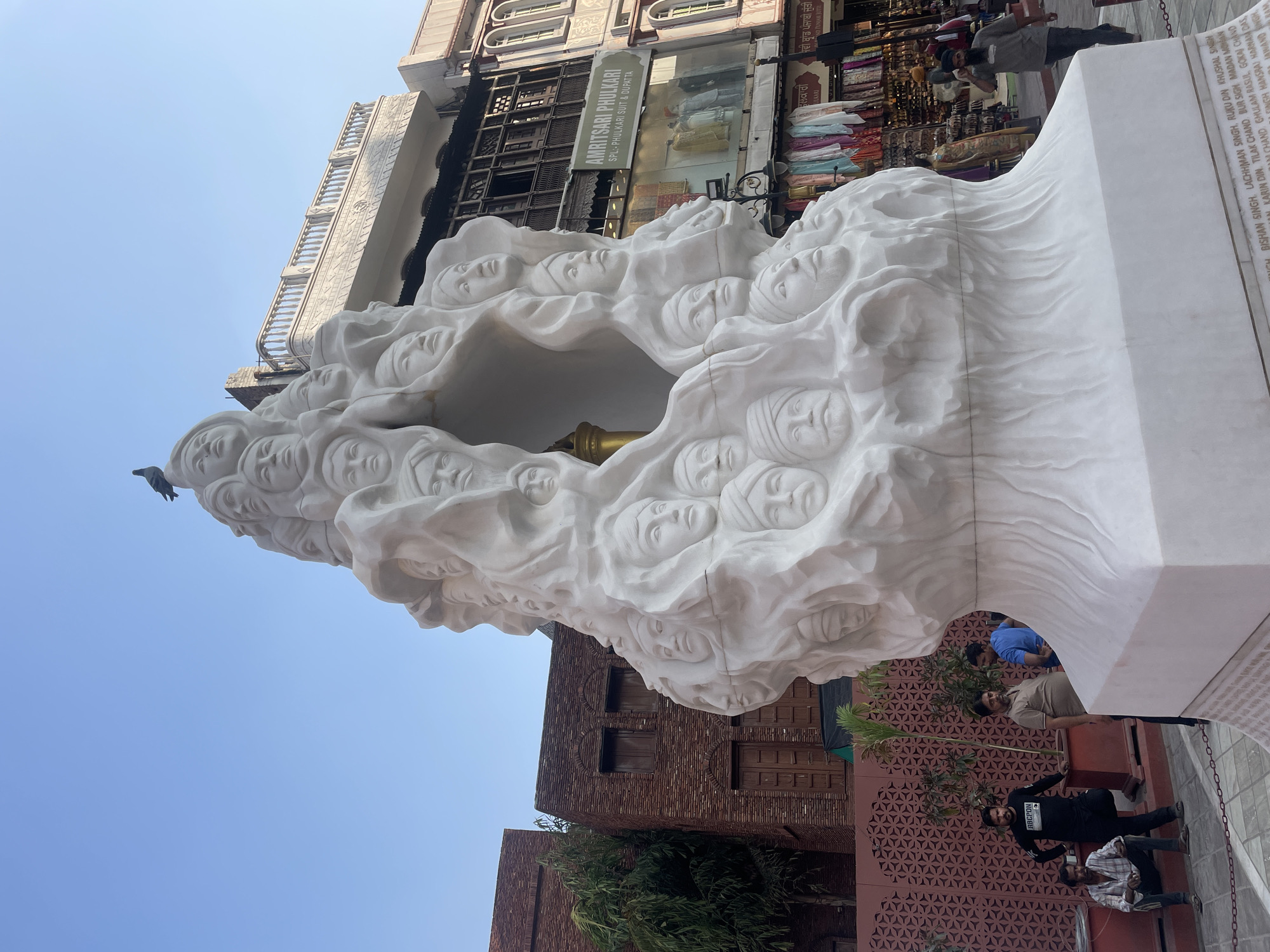 The image shows a large, white marble sculpture of many carved faces emerging from a flowing, abstract form. A pigeon is perched on top of the sculpture. In the background, there are brick buildings with wooden windows, a building with a balcony and signage that reads "AMRITSARI PHULKARI SPL: PHULKARI SUIT & DUPATTA", and shops with colorful textiles displayed. Several men are visible in the foreground and middle ground, some standing and some sitting. One man in the middle ground is walking and appears to be on his phone. The scene is outdoors, under a clear blue sky, and appears to be in a public area, likely a market or plaza in Amritsar, India, given the signage. The sculpture is the central focus, with people and the surrounding urban environment providing context.
