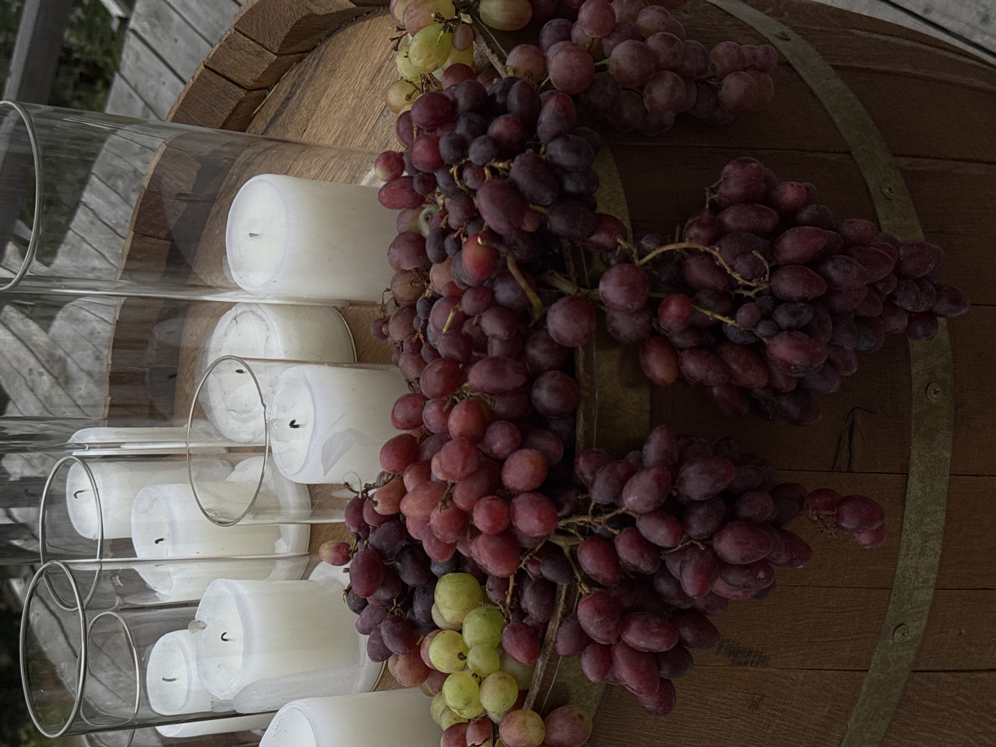 The image depicts a wooden barrel used as a decorative base for an arrangement of white pillar candles in glass cylinders and bunches of red and green grapes. The barrel, made of staves bound by metal bands, rests on a wooden surface that appears to be a deck or outdoor table, suggested by the visible wooden planks and railing in the background. The scene is set outdoors, with natural light illuminating the subjects. The composition is a close-up, focusing on the details of the barrel, candles, and grapes. The location is identified as Tibeta, Georgia.