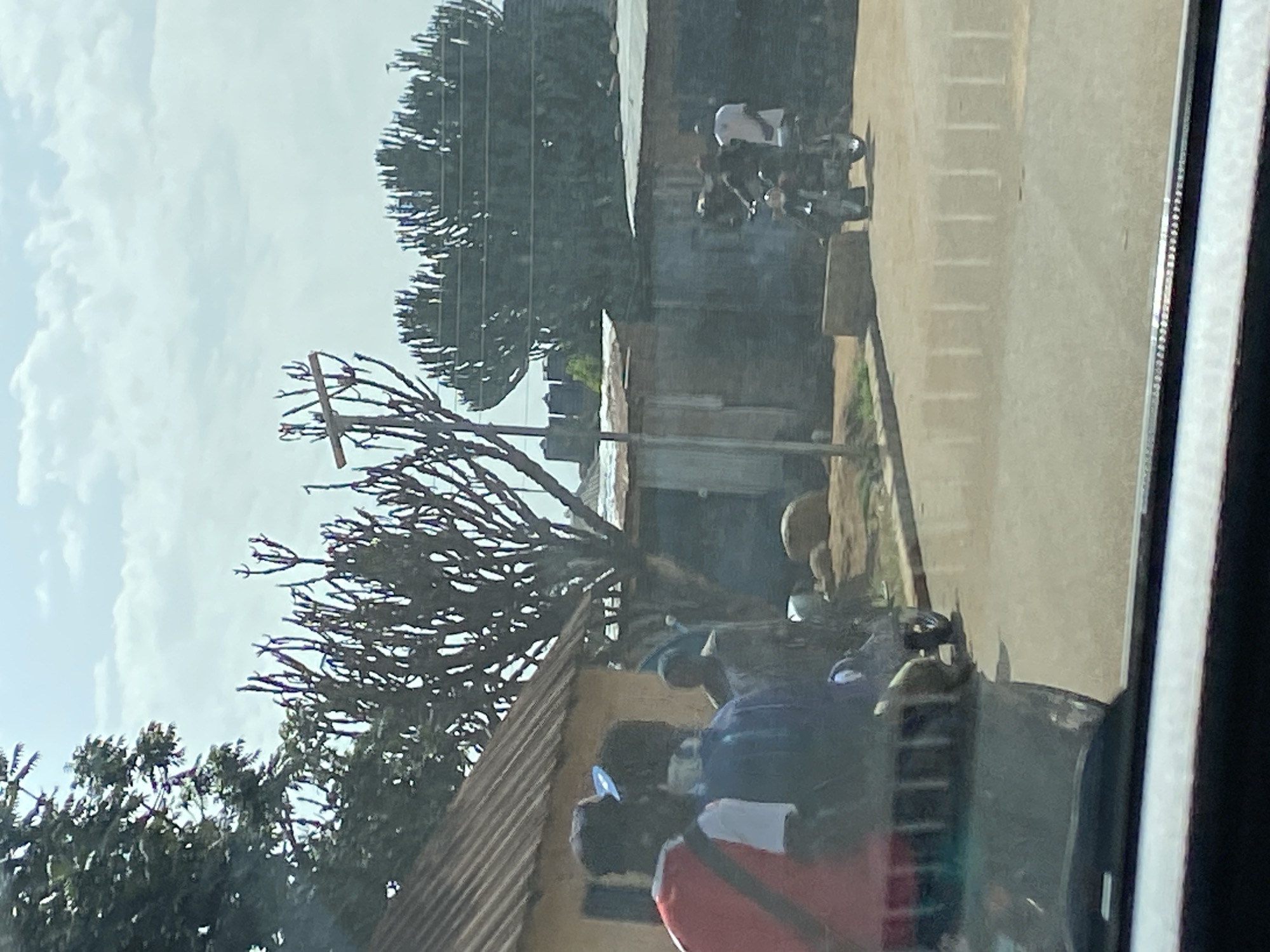 In Bar Jirgi Summa, Nigeria, a dusty, unpaved street is depicted on a bright, cloudy day. The foreground is dominated by the reflection of a vehicle's windshield, obscuring a clear view of the street's surface. In the middle ground, two individuals are seen from behind, one wearing a red shirt and the other a blue shirt. They are standing near a small, tan-colored building with a corrugated metal roof. Behind them, a leafless tree with branches resembling antlers stands tall. Further back, several structures constructed from corrugated metal and wood line the street, suggesting a densely populated or commercial area. Two men on a motorcycle are visible on the right side of the street, with one man standing beside it and the other seated on the vehicle. Smoke or dust is emanating from the rear of the motorcycle. In the background, large, mature trees with a distinctive branching pattern are present, and a few buildings can be seen in the distance. The overall impression is of a rural or semi-urban environment with modest infrastructure.