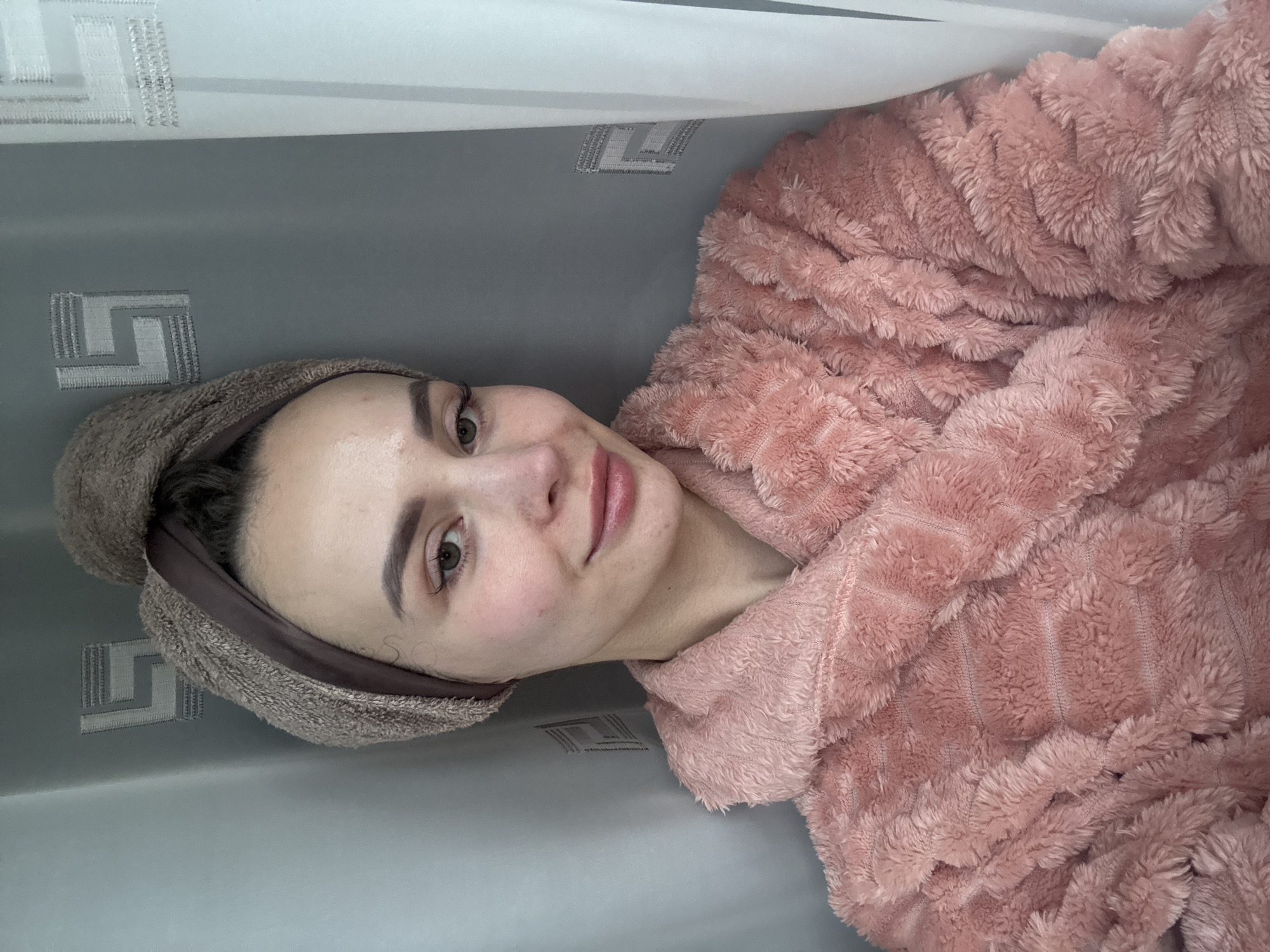 A woman is depicted in a close-up, chest-up shot, looking directly at the camera with a soft, pleasant smile. Her head is tilted slightly. She has light-colored eyes, well-groomed eyebrows, and naturally pink lips. Her skin appears clear.
She is wearing a voluminous, soft, and fluffy pink robe, possibly made of fleece or a similar textured fabric, which covers her shoulders and arms. Her hair is wrapped in a light brown, towel-like head wrap, suggesting she might have recently showered or is engaging in a self-care routine.
The background is simple, featuring a two-toned wall or curtain. The upper section is a plain light, possibly white or very light gray color. The lower section is a solid medium gray. On this grey background, a repeating geometric pattern, resembling a stylized Greek key or labyrinth motif, is visible. These patterns appear to be embroidered or woven into the fabric. The overall setting suggests an indoor environment, likely a home, in Kyiv, Ukraine. There is no discernible text visible in the image.