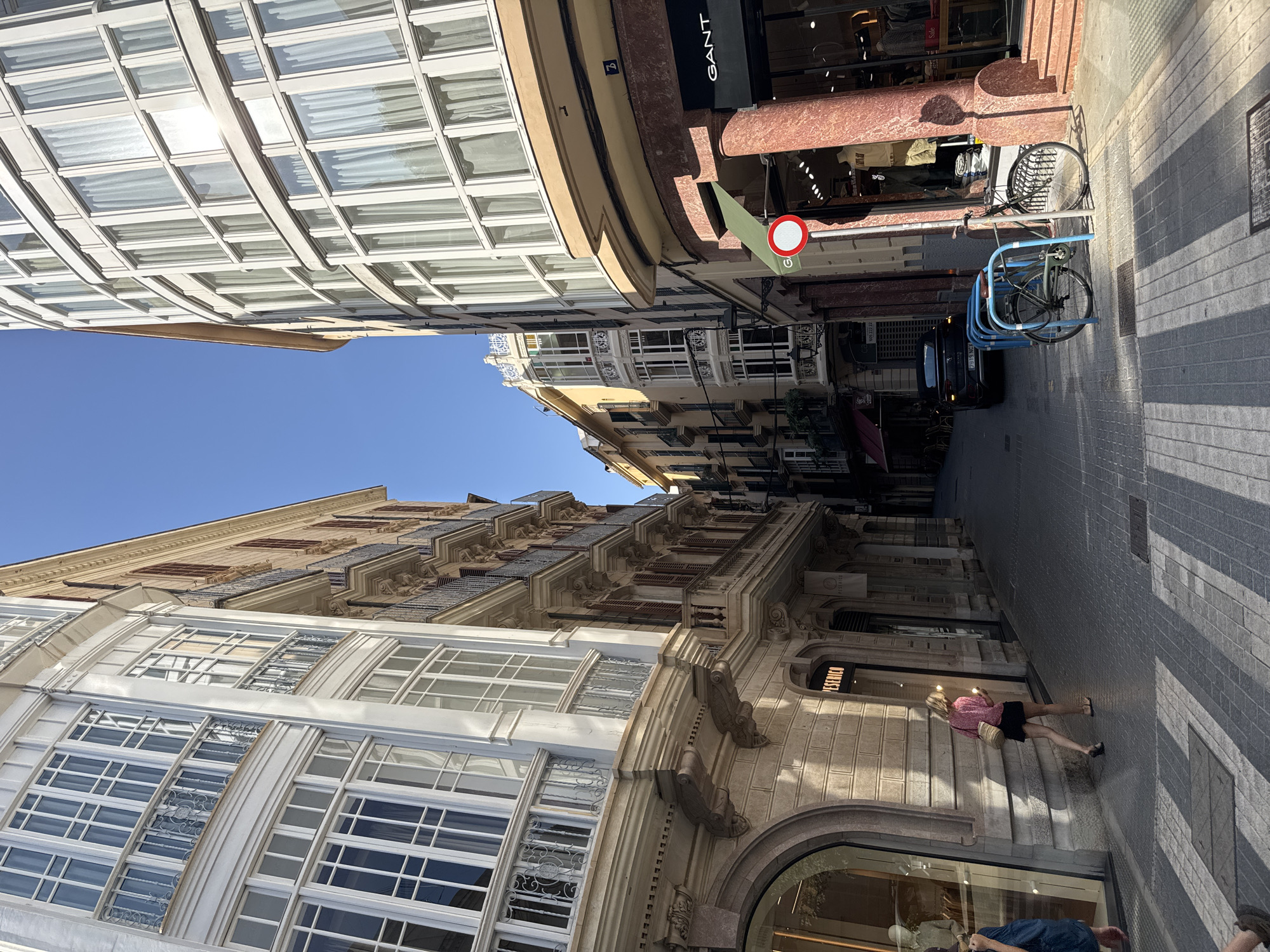 An eye-level, medium shot captures a sunlit narrow street in Palma, Spain, lined with historic buildings. On the left, a light-colored building features ornate white bay windows and balconies with intricate ironwork. Adjacent to it, a building with ochre-colored walls displays similar architectural elements. The right side of the street is dominated by a modern-looking building with a curved facade and numerous windows framed in white. This building's ground floor houses a Gant store, identifiable by its prominent signage and a display of clothing. A red and white circular sign is mounted on a pole near the store entrance. A bicycle is parked in a blue rack. The street is paved with grey cobblestones, and shadows from the buildings create a striped pattern. A black car is parked further down the street. A woman with blonde hair, wearing a pink top and black skirt, walks away from the camera, carrying a straw bag. Another person's legs are visible in the lower left corner of the frame. The sky above is a clear, bright blue.