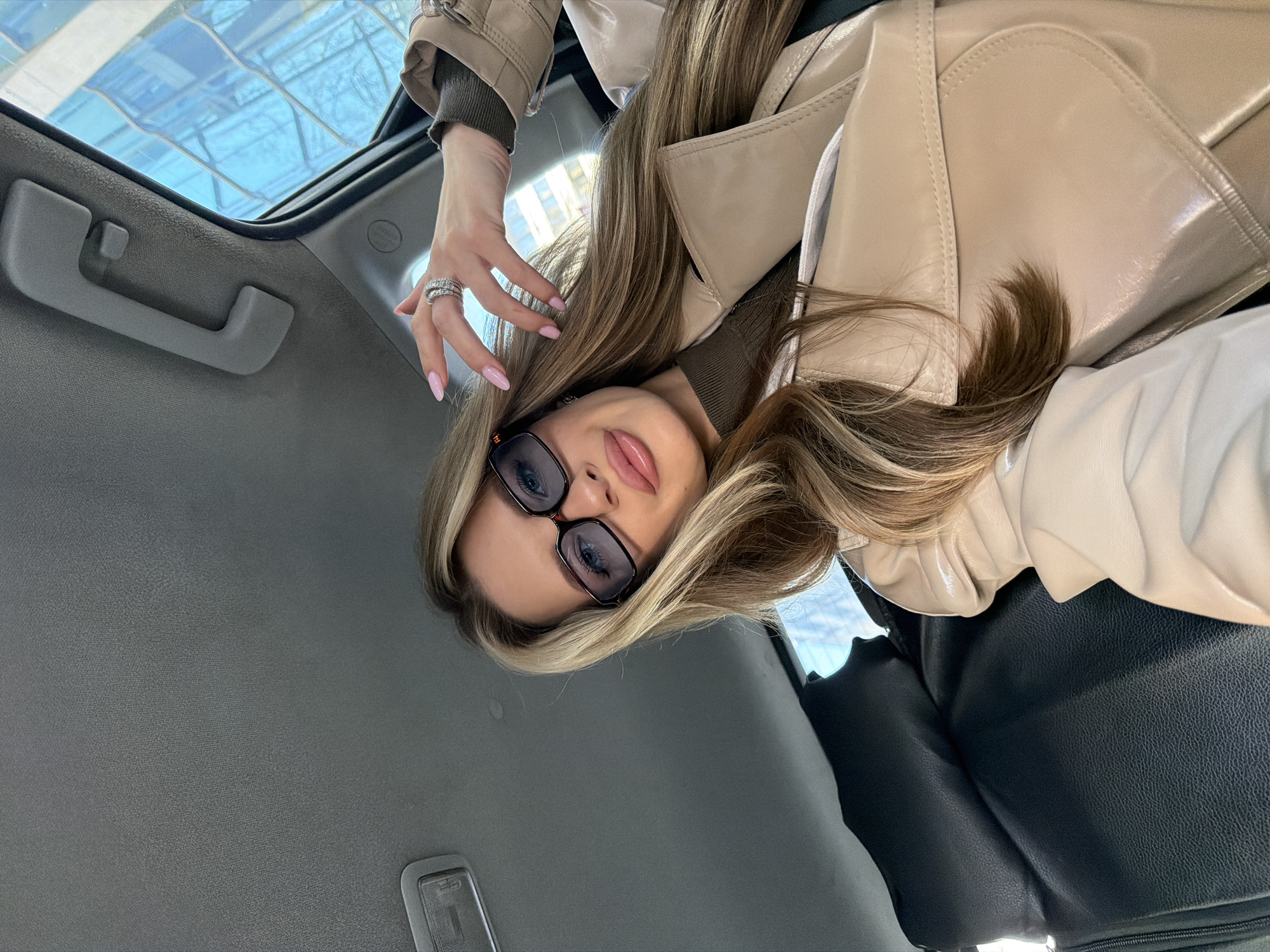 A young woman is in a car. She is wearing glasses, a beige coat, and jewelry. The car has a sunroof and a gray interior. Through the sunroof, a building with glass windows is visible. It appears to be a sunny day. The city is Berlin, Germany.