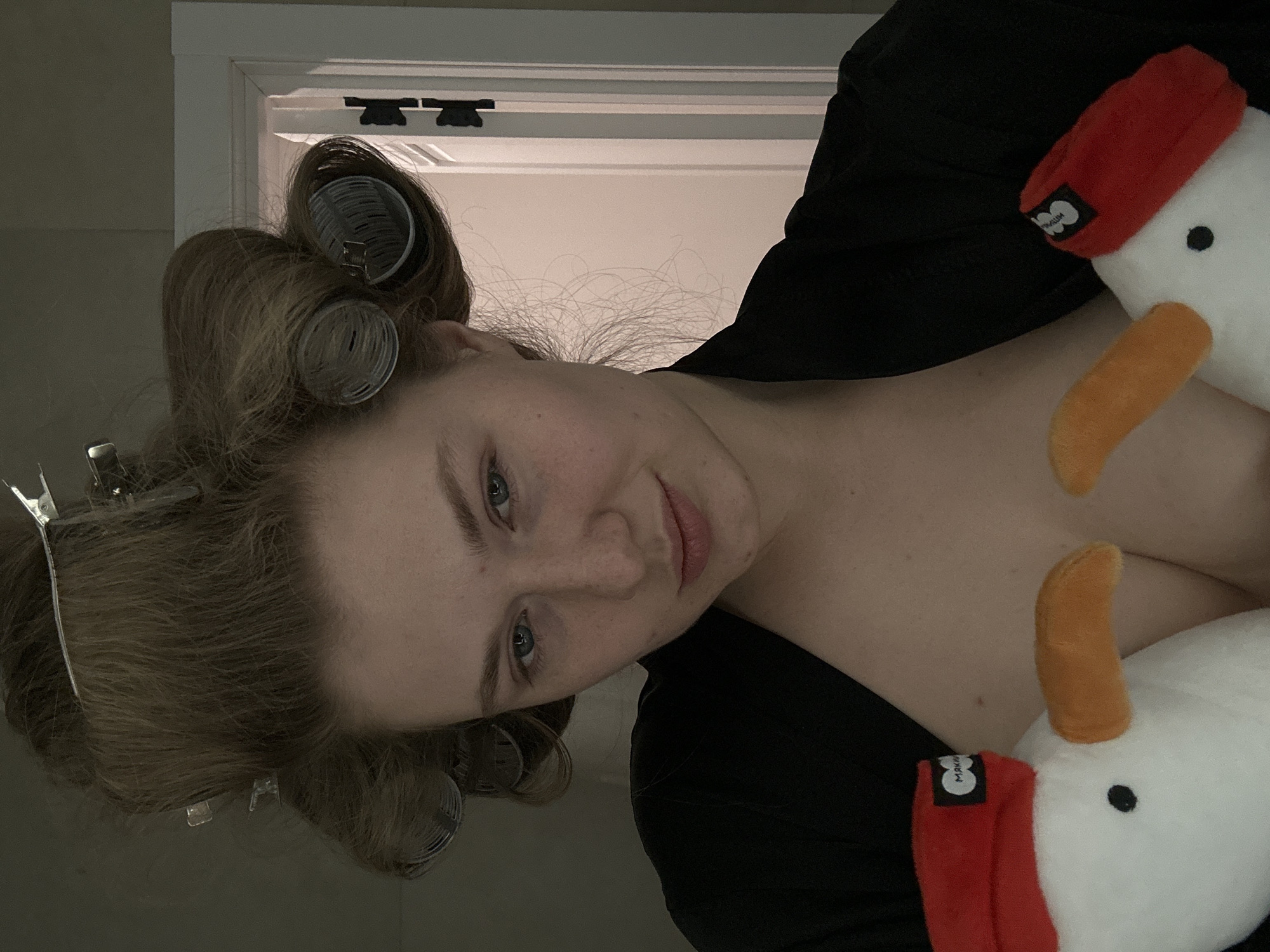 A woman is visible, wearing hair curlers and a black robe, cuddling two stuffed goose toys with red hats. The location cannot be determined from the image.