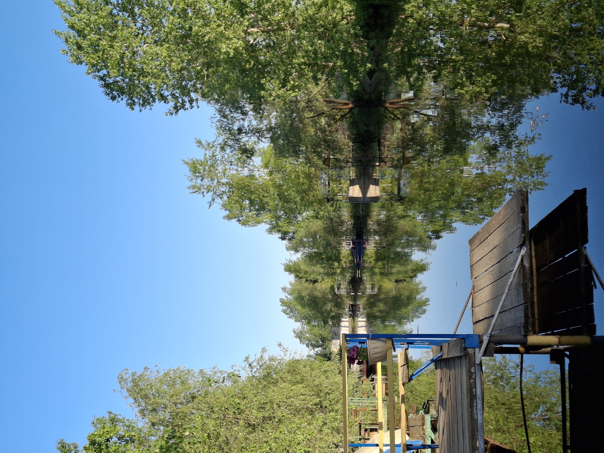 The image shows a scene in Chaikovsky, Russia. The view is taken from near the water's edge, looking toward a body of water, reflecting trees and the sky above. Various structures appear on the shoreline, including wooden planks, colorful metal railings, and supporting structures. Lush green vegetation lines the lower part of the scene. The reflection of trees and sky is visible in the water.