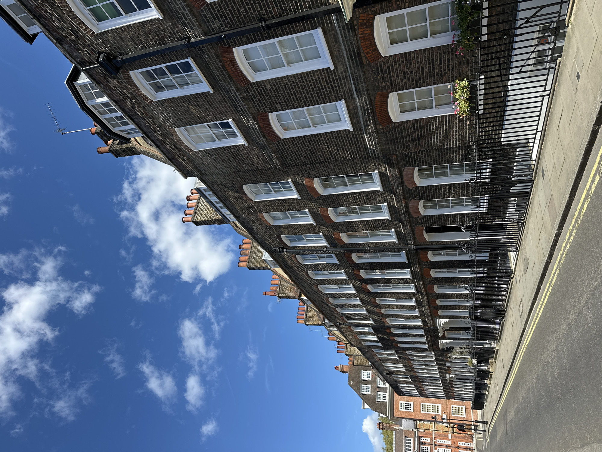The image captures a street scene in London, United Kingdom, featuring a row of traditional brick townhouses. These houses are characterized by their uniform architectural style, including arched windows with white frames. Black wrought-iron fences run along the sidewalk in front of the houses. A paved street is visible, marked with double yellow lines. The sky above is bright blue with scattered white clouds.