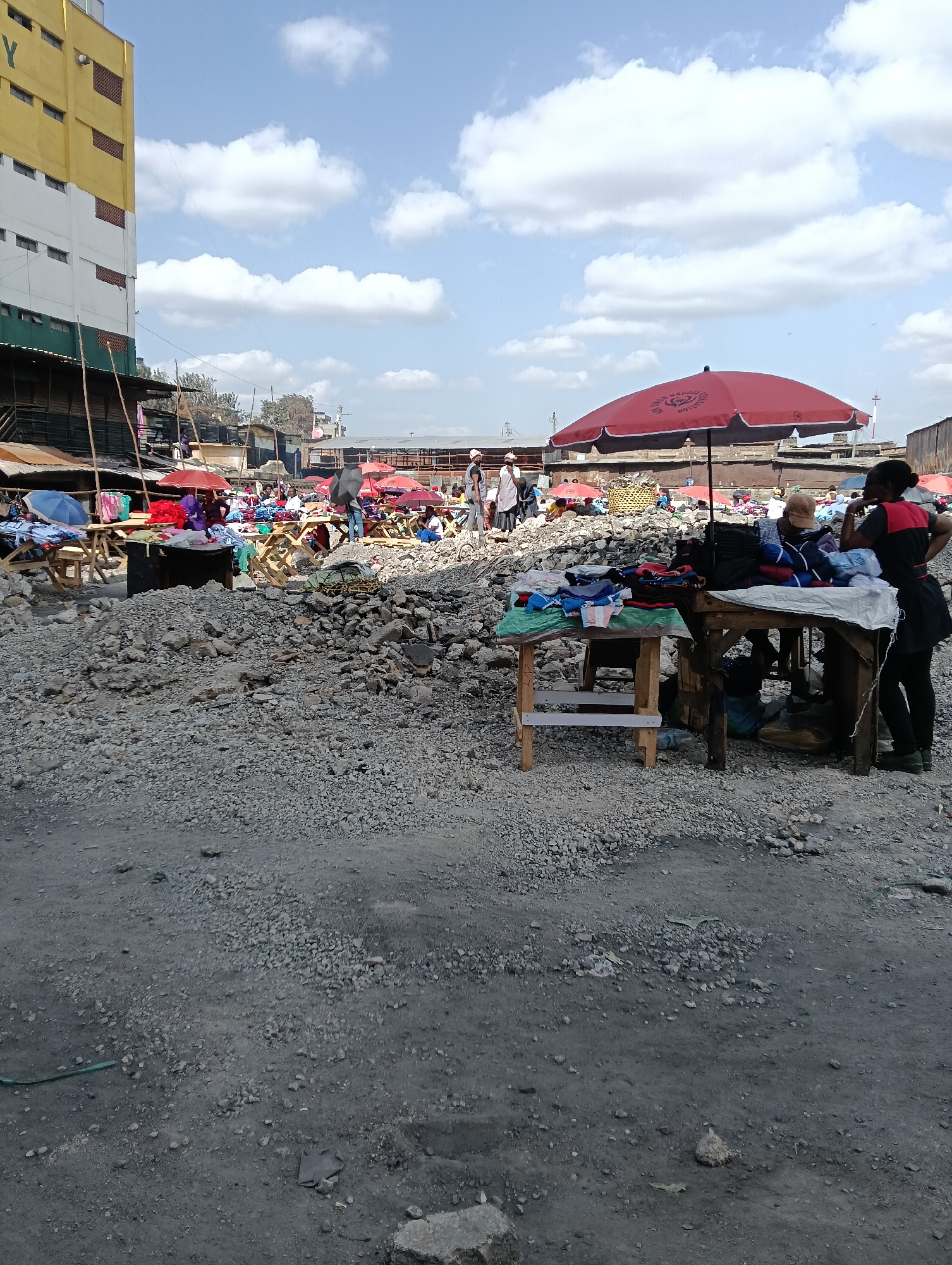 This media file captures an outdoor market scene in Nairobi, Kenya. The foreground is dominated by a rough, uneven ground covered in gravel and rubble. In the mid-ground, various market stalls are set up, featuring wooden tables displaying folded clothing. Brightly colored umbrellas, predominantly red, provide shade for the vendors and their merchandise.
Several people are visible throughout the scene, suggesting an active market day. Some individuals are tending to the stalls, while others appear to be browsing or moving through the market. The background shows a large, multi-story building with yellow and white coloring, indicating an urban setting. The sky is partly cloudy with scattered white clouds against a light blue backdrop, suggesting it is daytime, likely mid-morning or afternoon.
Notable details include the piles of rubble, which might indicate ongoing construction or a less-developed area within the city. The presence of umbrellas and the arrangement of goods on tables are typical of an open-air market. The clothing displayed on the tables varies in color and style. There is no visible text in the provided crops. The overall impression is one of a bustling marketplace with a gritty, utilitarian atmosphere.