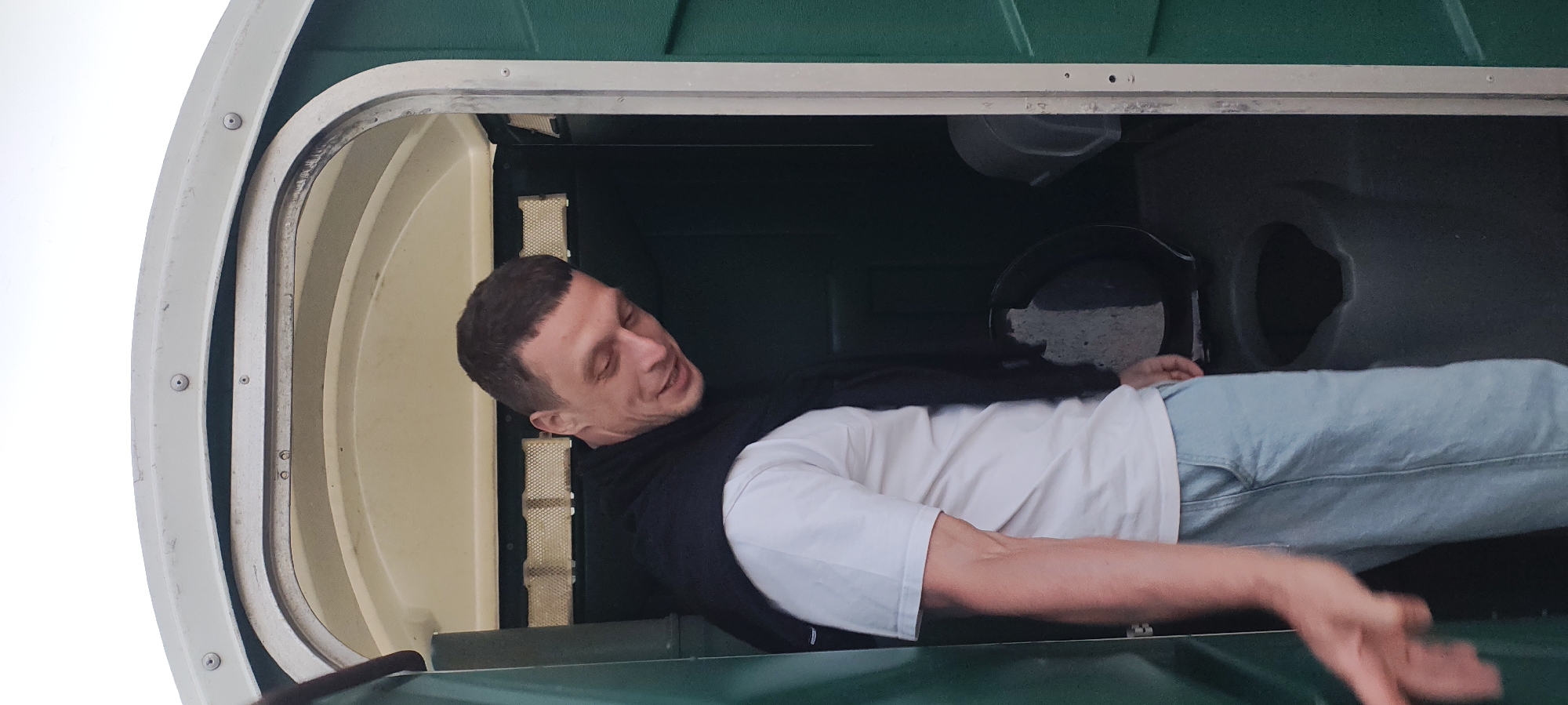 A man stands in the doorway of a portable toilet. He is wearing a white t-shirt, light blue jeans, and a black sweater draped over his shoulders. The man is looking down and to his right with a slight smile. His left arm is extended towards the camera. Inside the portable toilet, a dark-colored toilet is visible. The portable toilet is green. The scene appears to be outdoors, with a pale sky visible above the toilet.