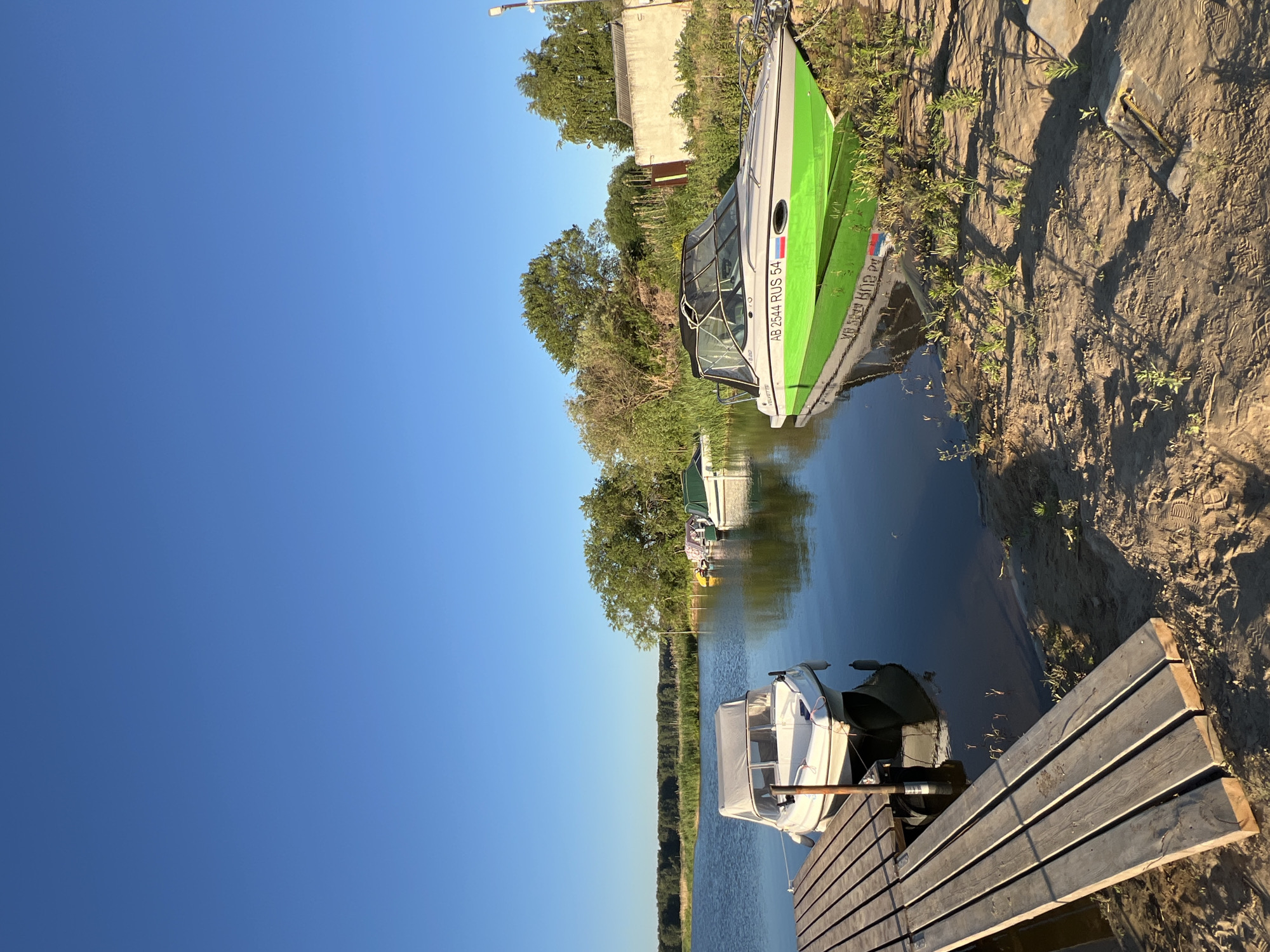 This is a full-frame, eye-level shot of a serene waterside scene in Novopichugovo, Russia. The clear blue sky dominates the upper half of the image. In the foreground, a wooden dock extends into the calm, reflective water. To the right of the dock, a white motorboat with a grey canopy is moored. Further in the background, several other boats, including a prominently featured green and grey speed boat with the registration "AB 2544 RUS 54", are visible. The boats are moored along a sandy bank with sparse vegetation. The bank is lined with trees and bushes, with a simple building structure visible in the far right background. The scene suggests a peaceful recreational area by the water. There are no people visible in the image.
