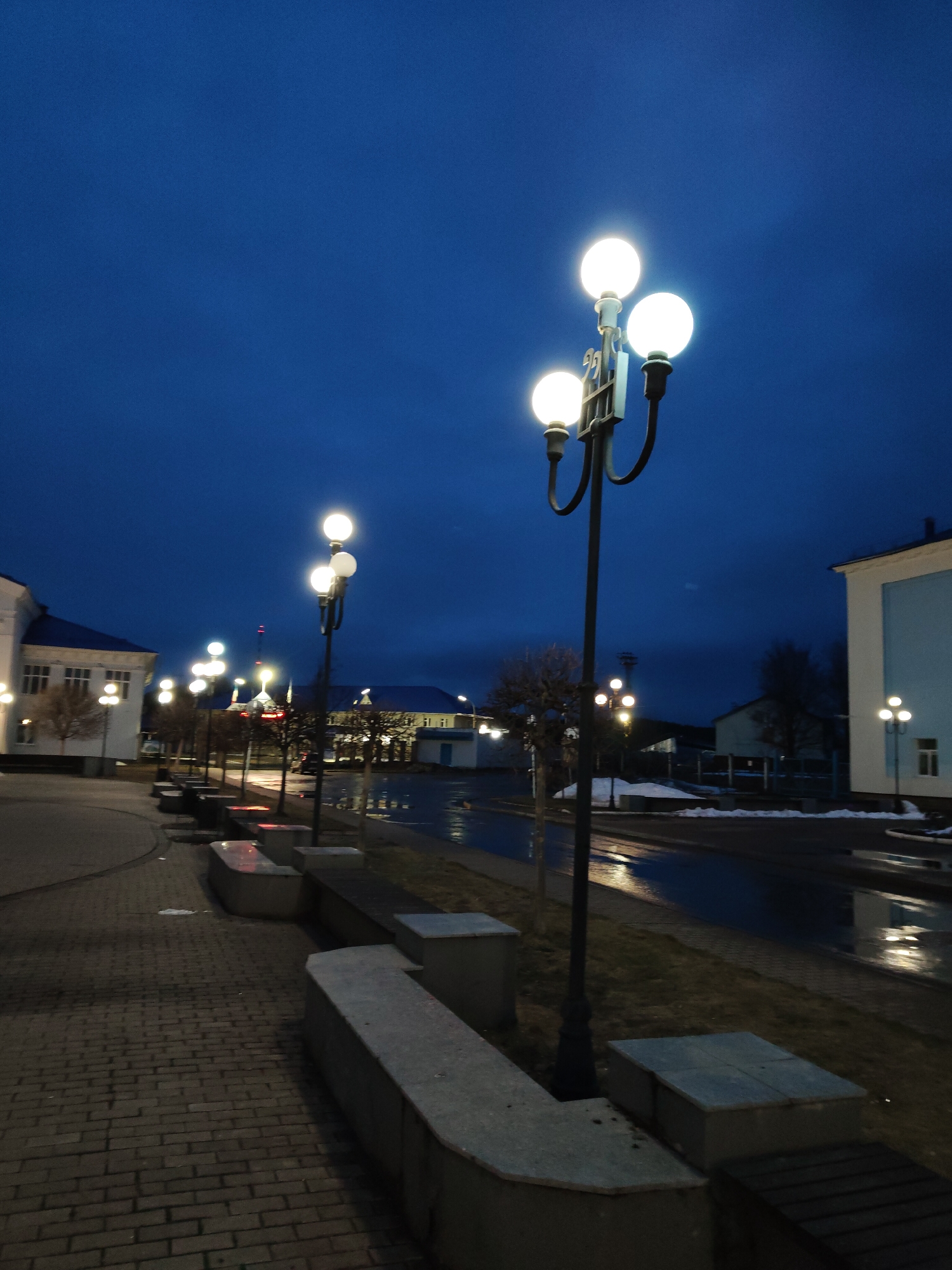 The image depicts a street scene in an unidentified city at dusk or night. The focal point is a row of ornate streetlights with three spherical bulbs each. These lights illuminate the scene, casting reflections on the wet pavement of the street. The sidewalk is made of brick and is lined with low, concrete benches. In the background, buildings can be seen, some illuminated, suggesting a small town or city center. The sky is a dark blue, indicating either late evening or pre-dawn. There are no people visible in the image. Patches of snow are also visible.