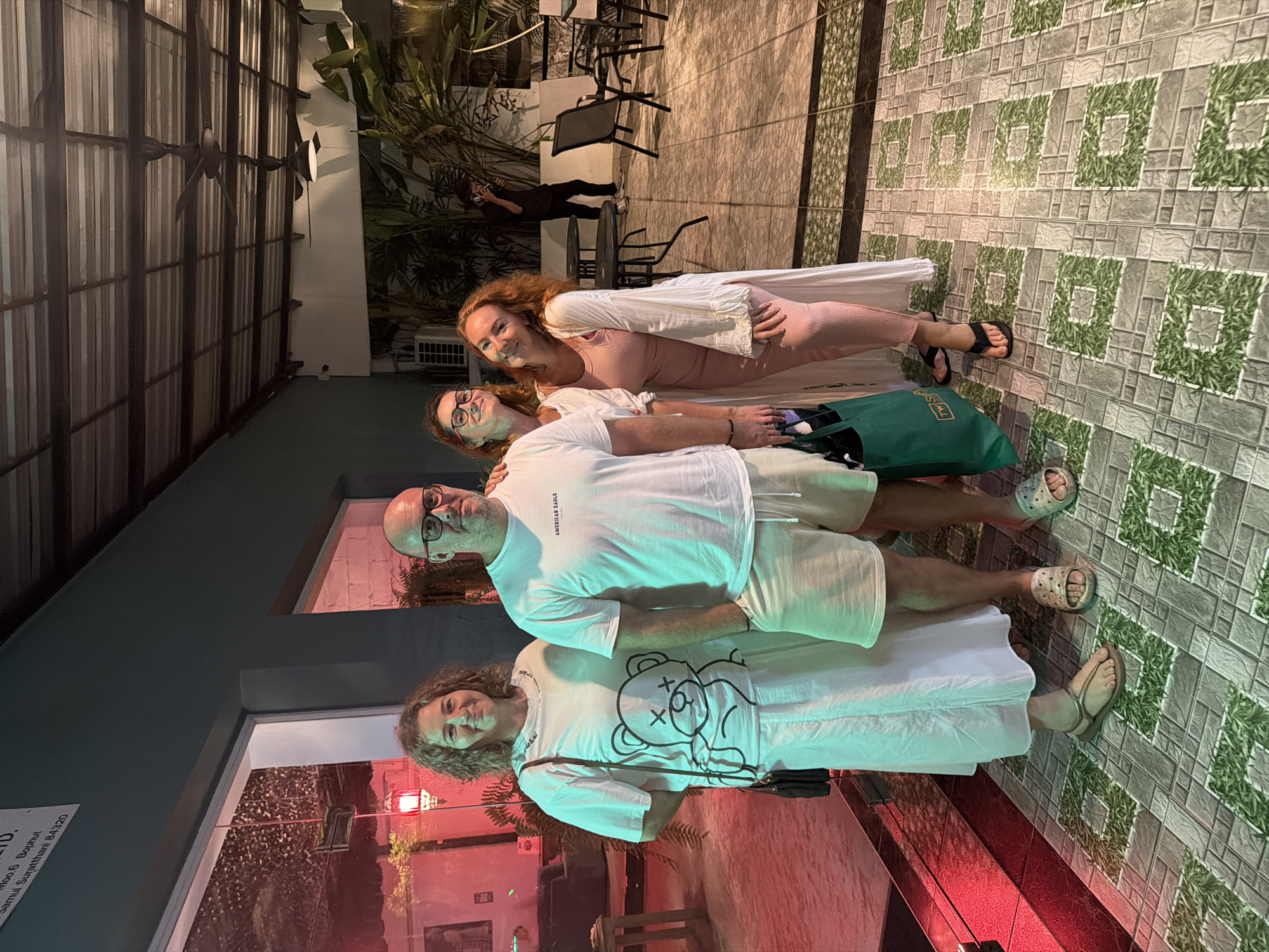 A group of four people are posing for a photo, with two couples standing in front of each other. The couple in the foreground consists of a man with glasses and a white t-shirt, and a woman with curly hair wearing a white t-shirt with a teddy bear graphic. Behind them, another woman with red hair is wearing a pink dress and a white cardigan, and a woman with glasses is wearing a white t-shirt. The background shows an outdoor setting with tiled flooring and some greenery, possibly a restaurant or bar area. The lighting suggests it might be evening or night. The overall mood seems relaxed and happy. There is some text visible on a sign in the background, but it is mostly unreadable. The location is identified as บ้านบางรักษ์, Thailand.