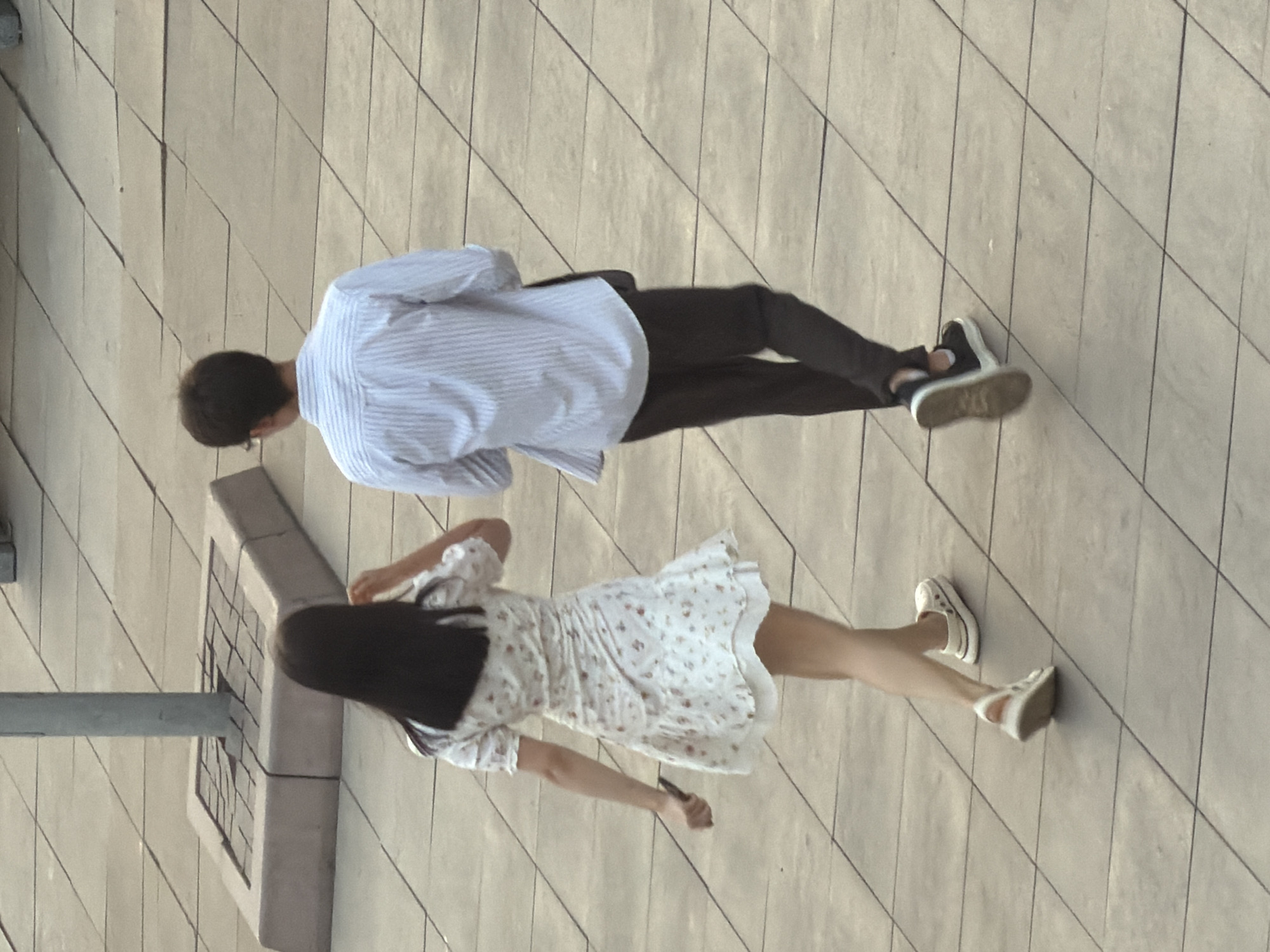 The media file shows two people walking on a tiled outdoor surface. The person on the left, facing away from the camera, is wearing a light blue and white striped long-sleeved shirt and dark pants. They are also wearing black and white sneakers. The person on the right, also facing away, is wearing a white dress with a floral pattern and white sneakers. Their left arm is extended, and they are holding a dark object, possibly a phone or a small bag.
The scene appears to be outdoors during the daytime, with the lighting suggesting it's a bright day. The tiled surface is made up of large, rectangular tiles arranged diagonally. In the background, there is a concrete structure with a grid pattern on its top surface. The overall setting appears to be an urban or public outdoor space. There is no visible text in the image. The image is taken from a high angle, looking down on the two individuals. The people are walking away from the camera, so their facial expressions are not visible.