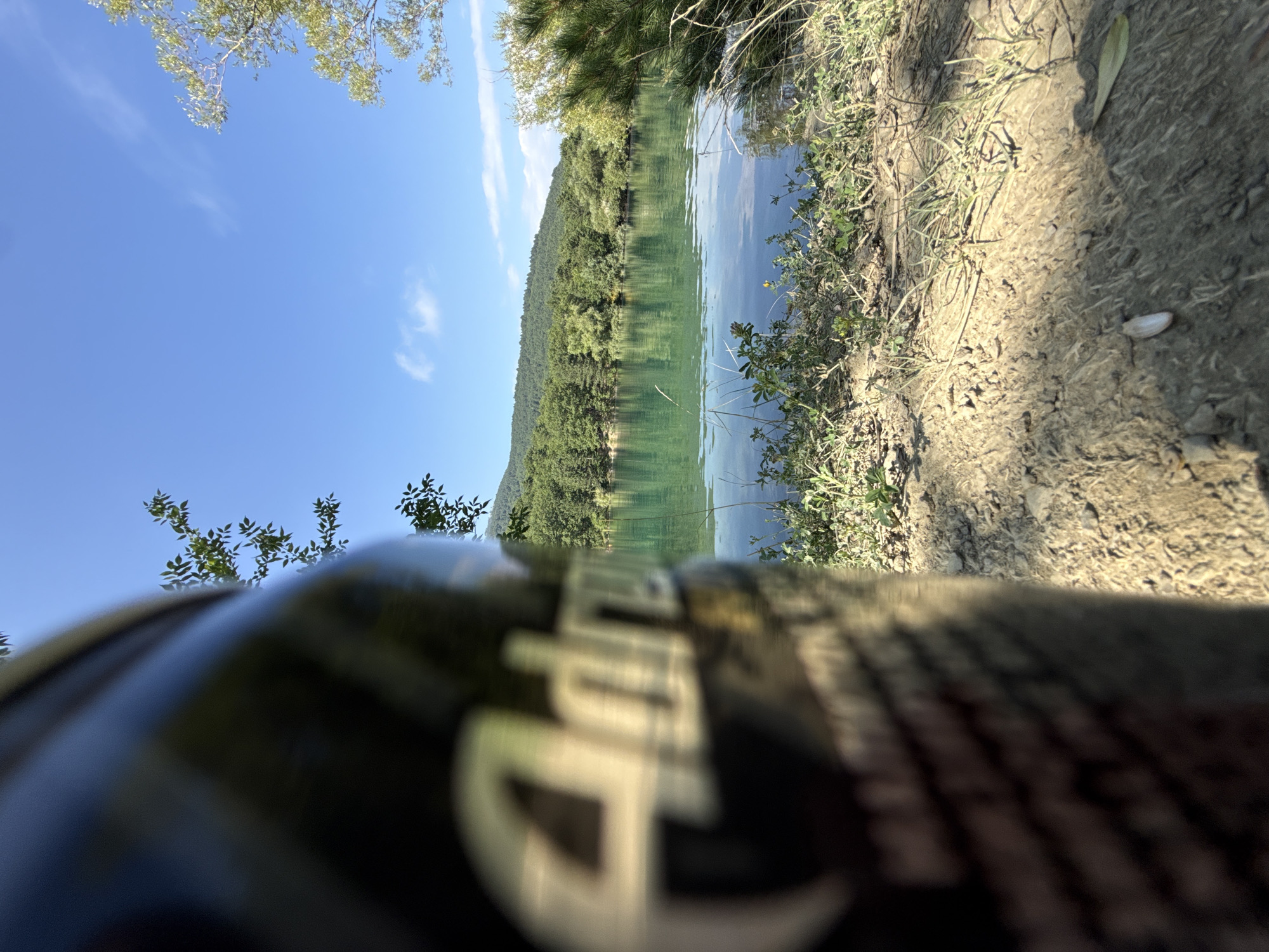 In the foreground, a dark, blurry object, possibly a container, occupies the left side of the frame, with partial text that appears to read "Adria." Beyond this, the scene opens to a picturesque natural landscape under a clear blue sky with scattered white clouds. A tranquil, turquoise lake stretches across the middle ground, its surface reflecting the surrounding lush green trees and a distant, tree-covered hillside. The shoreline is lined with dry grasses and some green vegetation. The overall impression is one of a serene outdoor setting. There are no people or animals visible in the image. The location is described as the city of Unknown, Ukraine.