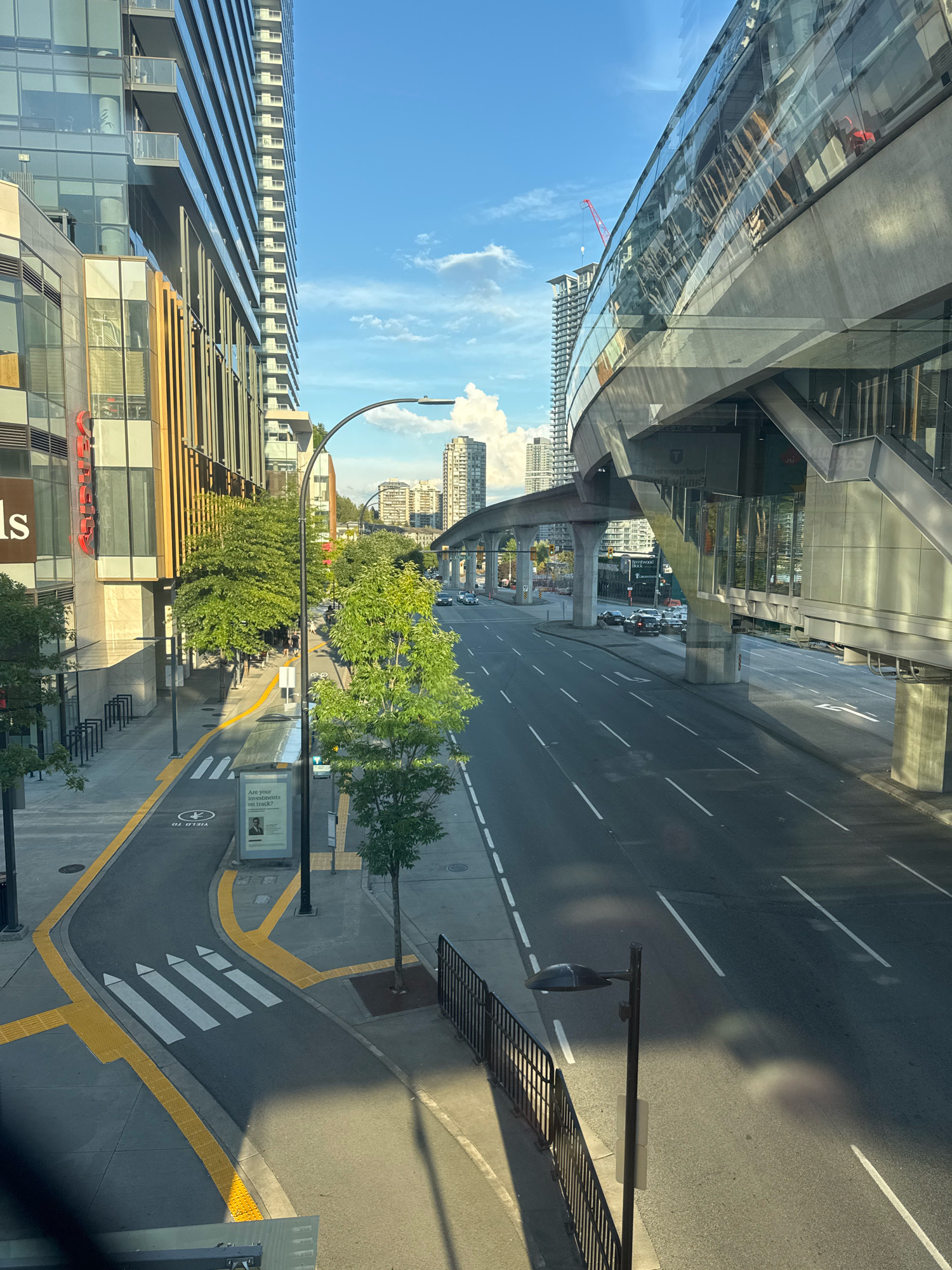 The image depicts a street scene in Burnaby, Canada, near a SkyTrain station. On the left, a modern building with glass and wood facade stands tall, with balconies and windows reflecting the sky. Adjacent to it, a paved sidewalk runs alongside the building, with bike racks and streetlights. A marked crosswalk with yellow tactile paving leads to the sidewalk. Further down the street, a lush green tree stands by the sidewalk.
The street itself is a multi-lane road with white lane markings. On the right side of the road, a modern SkyTrain station structure with a sleek, curved glass facade is elevated above the street, supported by concrete pillars. A pedestrian walkway with stairs leads up to the station platform. In the distance, more buildings and trees can be seen, under a clear blue sky with scattered white clouds. A red construction crane is visible in the background. Several cars are driving on the road. A streetlight with a modern design casts a shadow on the road. The overall impression is one of an urban environment with contemporary architecture and public transportation infrastructure.