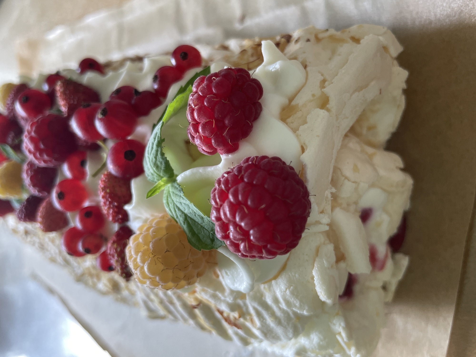 A close-up, overhead shot shows a dessert resembling a pavlova roll, topped with fresh berries and whipped cream. The dessert is adorned with raspberries, red currants, and what appear to be wild strawberries, along with a few sprigs of mint. The texture of the meringue is visible, with its characteristic crisp exterior and soft interior. The dessert is placed on a parchment paper, which is on a light brown surface, possibly a table. There are no people or any contextual information about the location in Дресвяново, Russia.