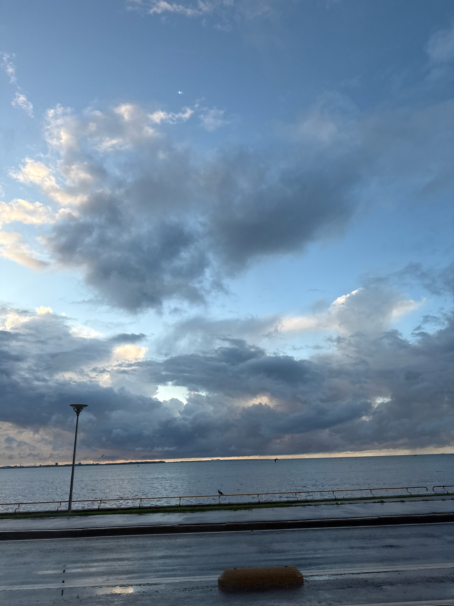 The image depicts a coastal scene with a cloudy sky over the sea. In the foreground, there is a wet asphalt road and a sidewalk. A lamppost stands on the sidewalk, and a yellow railing separates the sidewalk from a grassy area. The sea stretches to the horizon, where a city skyline is visible in the distance. The sky is filled with dramatic, dark gray and white clouds, with patches of blue sky peeking through. A small crescent moon is visible in the upper portion of the sky. There are a few small boats or buoys on the water. The overall atmosphere suggests it may be dawn or dusk, with the weather appearing overcast and potentially stormy. The location is identified as Tallinn, Estonia.