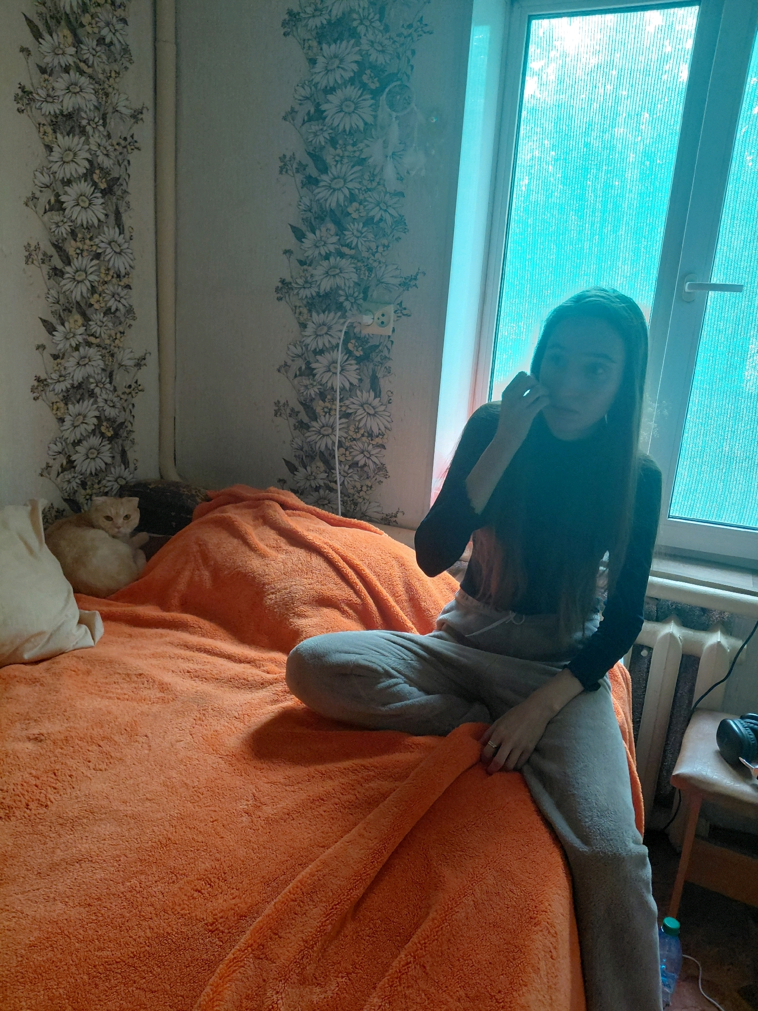 A young woman with long dark hair sits on a bed covered with an orange blanket. She is wearing a black long-sleeved shirt and grey sweatpants. Her left hand is near her face, and she appears to be touching her cheek. To the left of the woman, a light-colored cat is curled up on the bed. In the background, there is a window with blue-tinted glass, and the walls are covered with floral wallpaper. A radiator is visible near the window. The scene appears to be indoors, likely a bedroom, and the lighting suggests it could be daytime, though the blue tint of the window might obscure the true time of day.