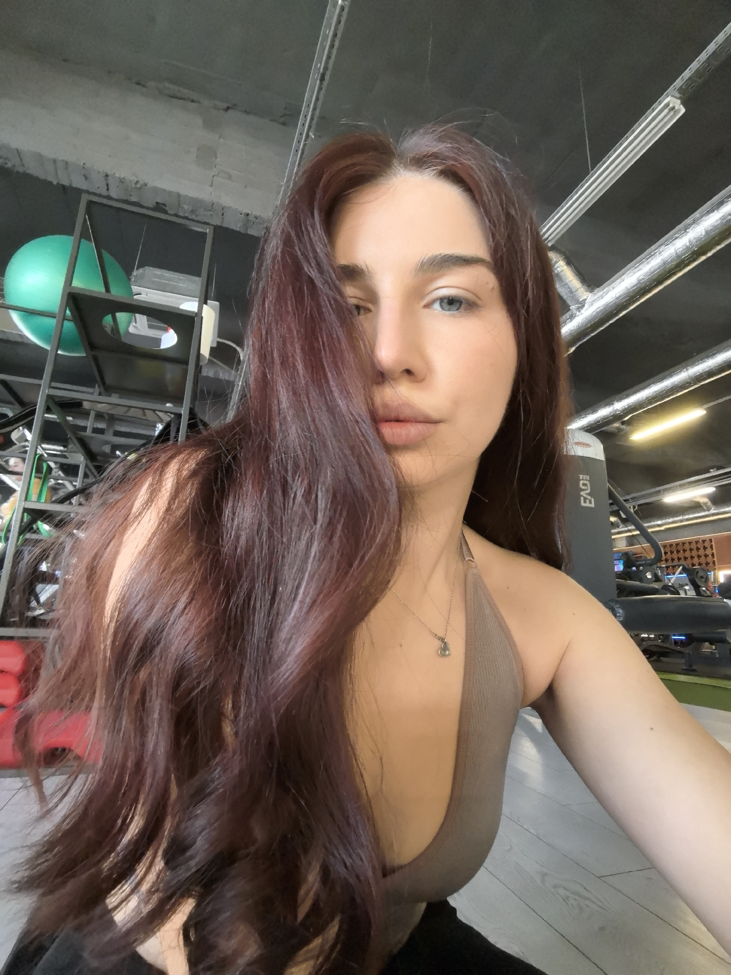 A woman with long, wavy auburn hair stands in a gym in Stara Zagora, Bulgaria. She is fair-skinned with blue eyes and is wearing a brown halter top and a delicate necklace. Part of her hair covers the left side of her face. The gym environment is visible in the background, featuring exercise equipment, a green exercise ball on a rack, exposed pipes on the ceiling, and a gray floor. The image appears to be a selfie taken at a close-up angle.