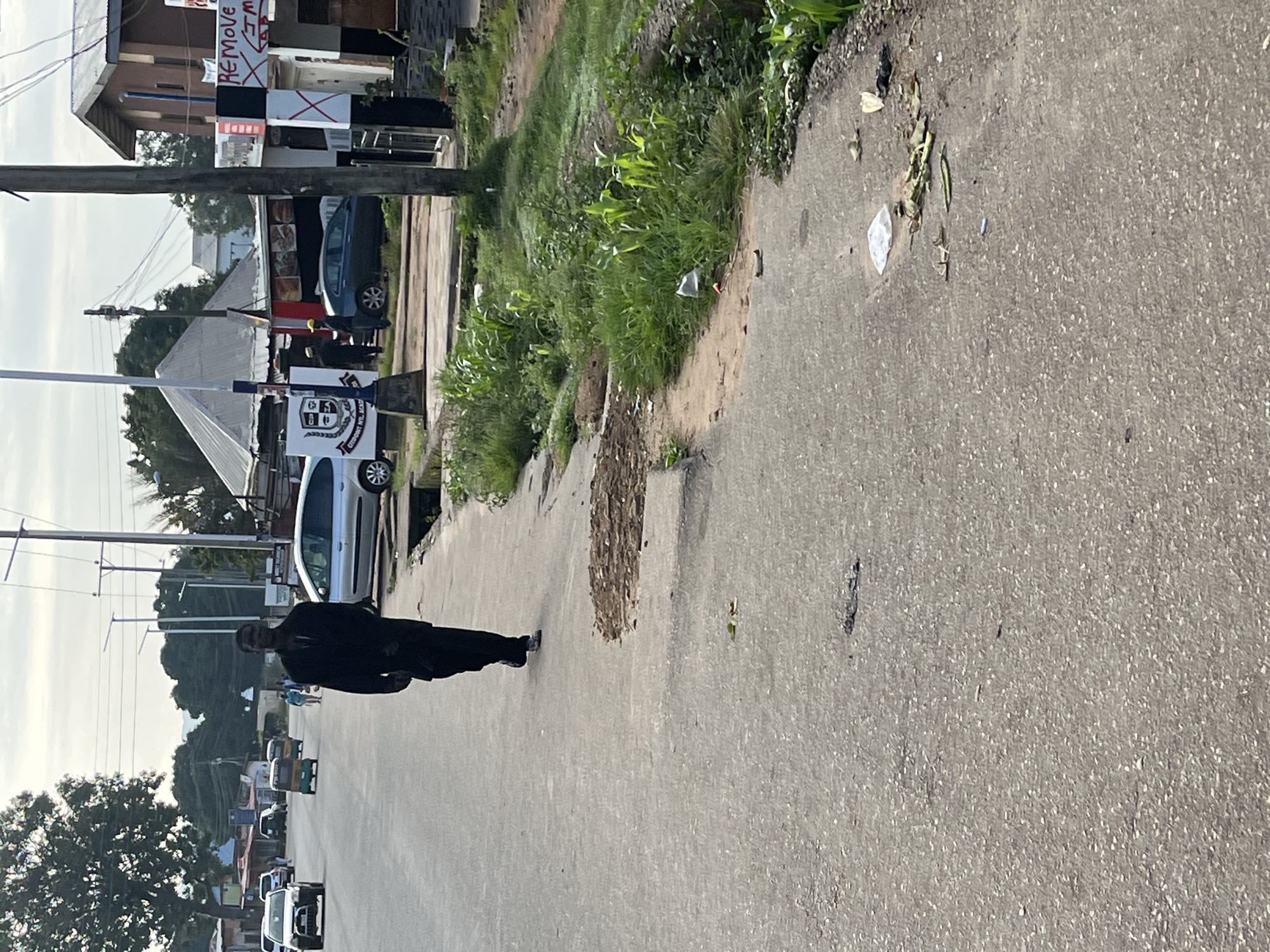 A medium shot captured on a slightly overcast day shows a man walking towards the camera on a paved road in Jos, Nigeria. The man, dressed in dark clothing, walks with his head slightly bowed. The road appears to be a two-lane street with a rough, worn surface. To the right of the road, a grassy embankment with scattered debris and some vegetation slopes upward. Further to the right, buildings and shops line the street. One building has a prominent sign with red and white markings. In the background, several vehicles, including cars and a tricycle, are visible, along with utility poles and wires. Trees are scattered throughout the scene. The overall impression is of a typical urban street in Jos, with a mix of residential and commercial structures and the presence of daily life activities.