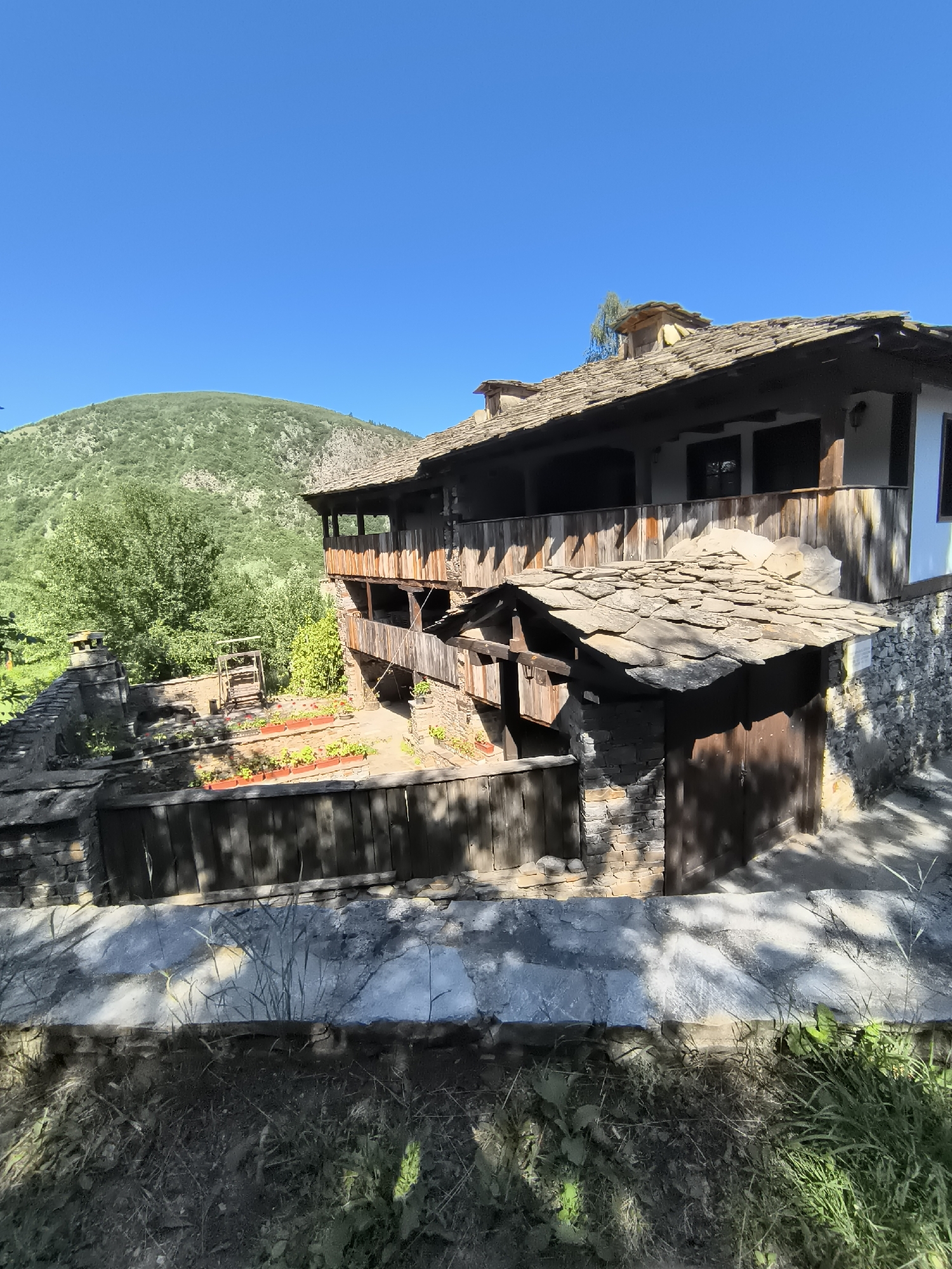 The image depicts a traditional Bulgarian house in the architectural reserve of Kovachevitsa, Bulgaria. The house is characterized by its stone and wooden construction, with a prominent stone foundation and walls, and a roof made of stone slabs. A wooden balcony with railings extends across the upper level. In the foreground, there is a wooden fence and a small garden with red flowers in planters. Behind the house, a lush green hill rises under a clear blue sky. The overall scene conveys a sense of rustic charm and historical preservation.