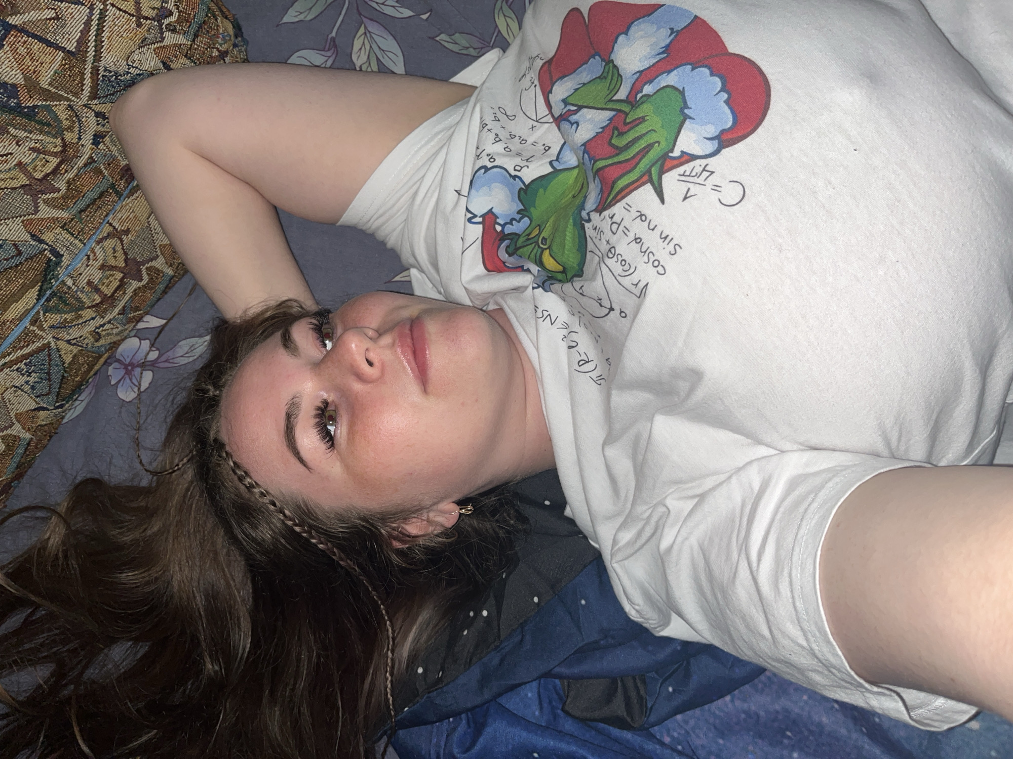 A young woman lies on her back on a patterned surface, possibly a bed or couch. She wears a white t-shirt with an image of the Grinch and handwritten formulas. Her brown hair is spread around her head, with small braids visible on one side. She has fair skin, and her eyes are open. The background is a mix of patterned fabrics and possibly some bedding. The location (Chaikovsky, Russia) cannot be determined from the image.