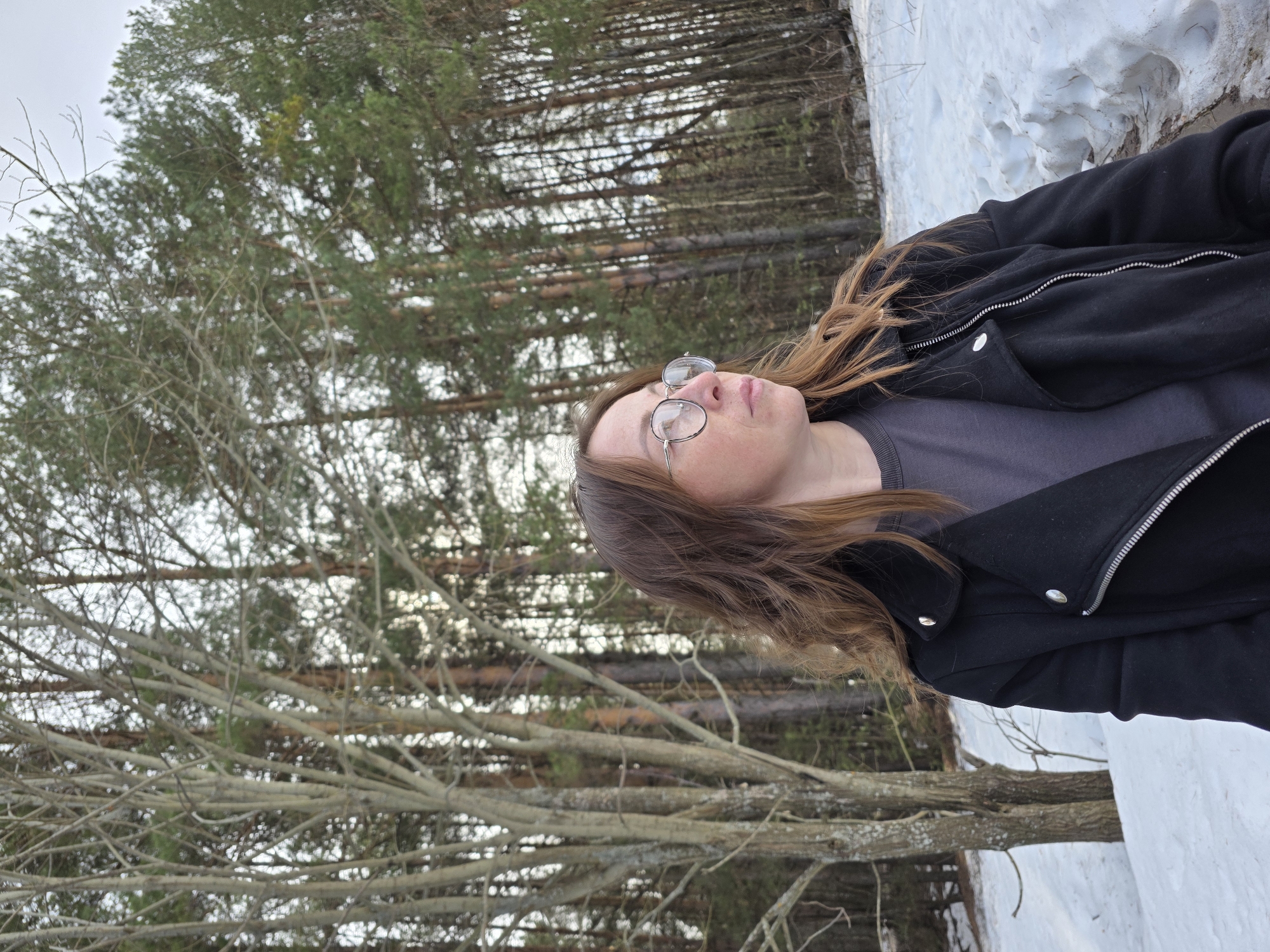 A person with long, light brown hair and glasses is lying in the snow. They are wearing a black jacket and a gray shirt. The person is looking up at the trees in the background. The location is Chaikovsky, Russia, as indicated in the prompt. The environment includes snow on the ground and a forest of trees in the background, some with green leaves and others with bare branches. The scene appears to be taken outdoors during the daytime.