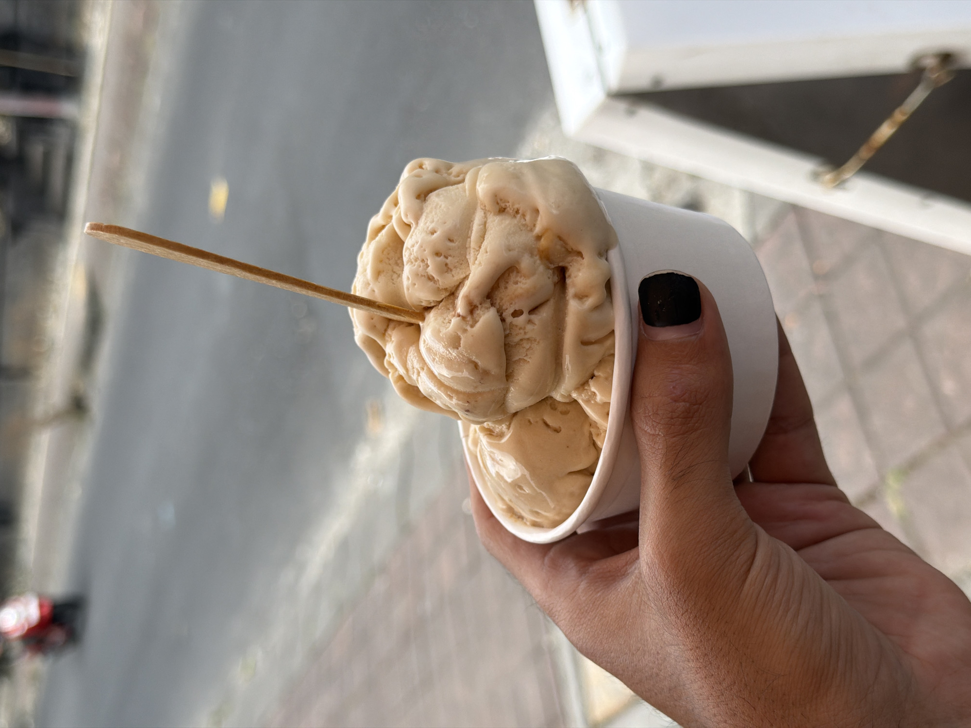 A close-up shot captures a hand holding a white paper cup filled with two scoops of light brown ice cream, with a small wooden stick inserted into the top scoop. The ice cream appears to be melting slightly. The background is blurred, showing a street scene with a red motorcycle in the distance and a light-colored object on the right. The scene is set on a sidewalk in North Kuta, Indonesia. The hand has visible veins and hair, and a black nail polish is visible on one finger.