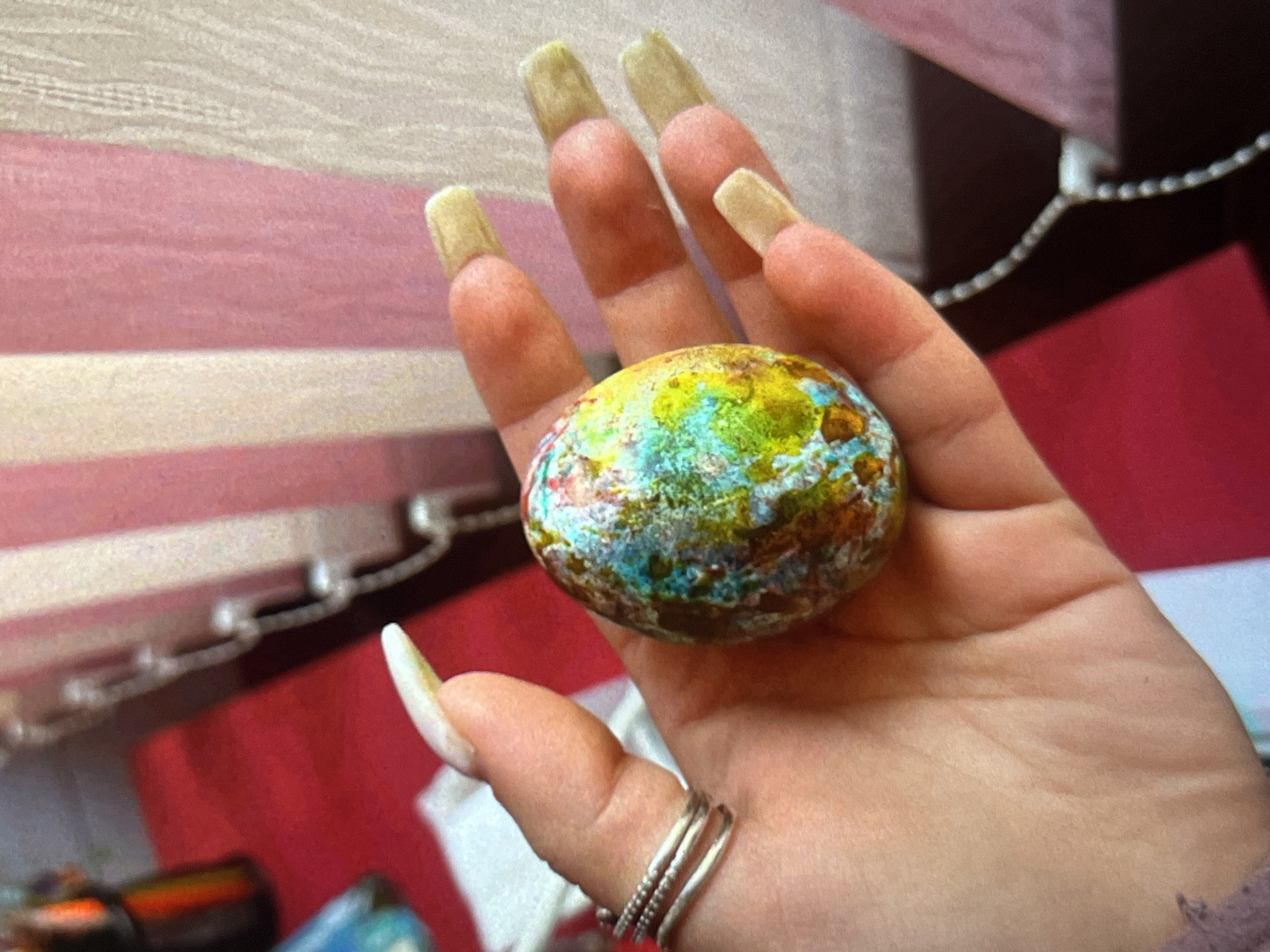 A hand, adorned with long, artificial fingernails, holds a multi-colored, egg-shaped object. The fingernails appear rectangular at the tip and are colored in a pale, yellowish hue. The object held in the palm is speckled with blue, green, brown, and yellow pigments. Three rings are visible on the thumb of the hand. The background consists of a blurred mix of pink and white horizontal blinds. It is not possible to infer any location in Plovdiv, Bulgaria from the provided information.