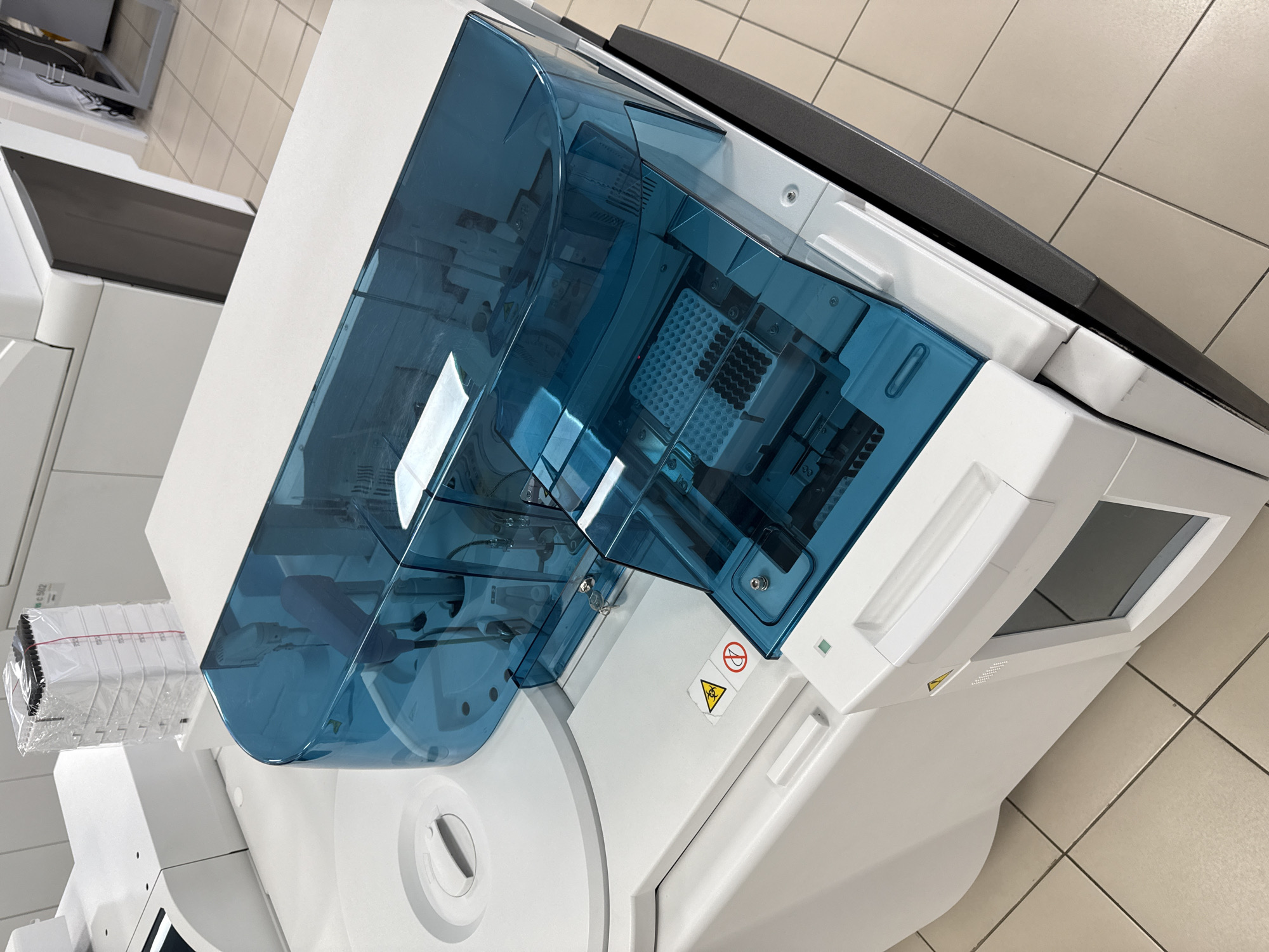 A clinical chemistry analyzer sits on a tiled floor. The analyzer is white with a translucent blue hinged cover. Inside, multiple small vials are visible, arranged in a grid pattern. A biohazard symbol is displayed on the machine's side panel. A stack of lab supplies is on the left side of the machine.