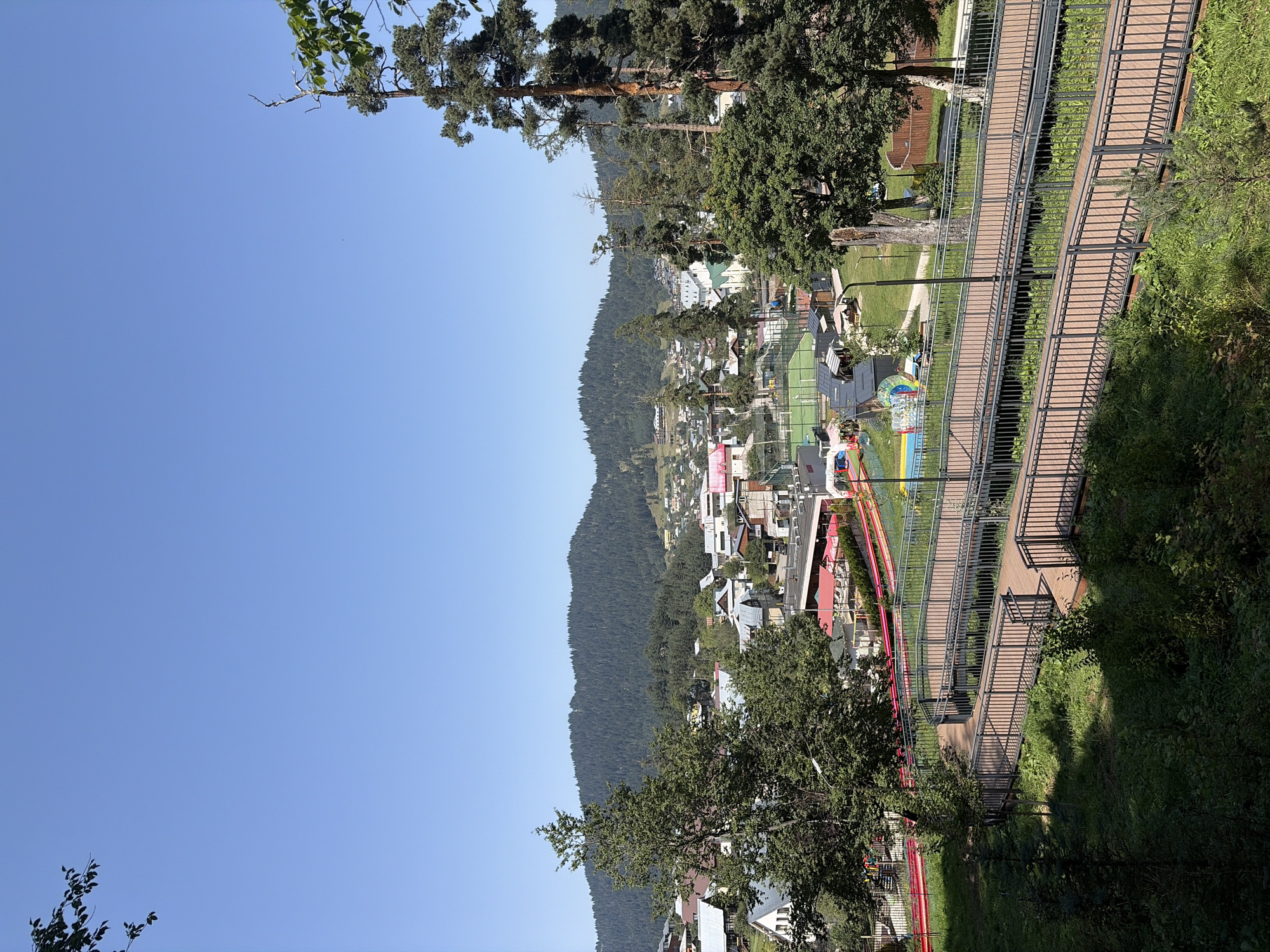 The image depicts a panoramic view of Bakuriani, Georgia, on a clear, sunny day. In the foreground, a modern wooden walkway with metal railings winds through lush green vegetation. Beyond the walkway, a resort or village area is visible, characterized by a mix of houses with gabled roofs and larger buildings with red awnings. A sports complex with a green field and a running track is also present. In the background, a densely forested mountain range rises against a bright blue sky. The architecture suggests a mountain resort town.