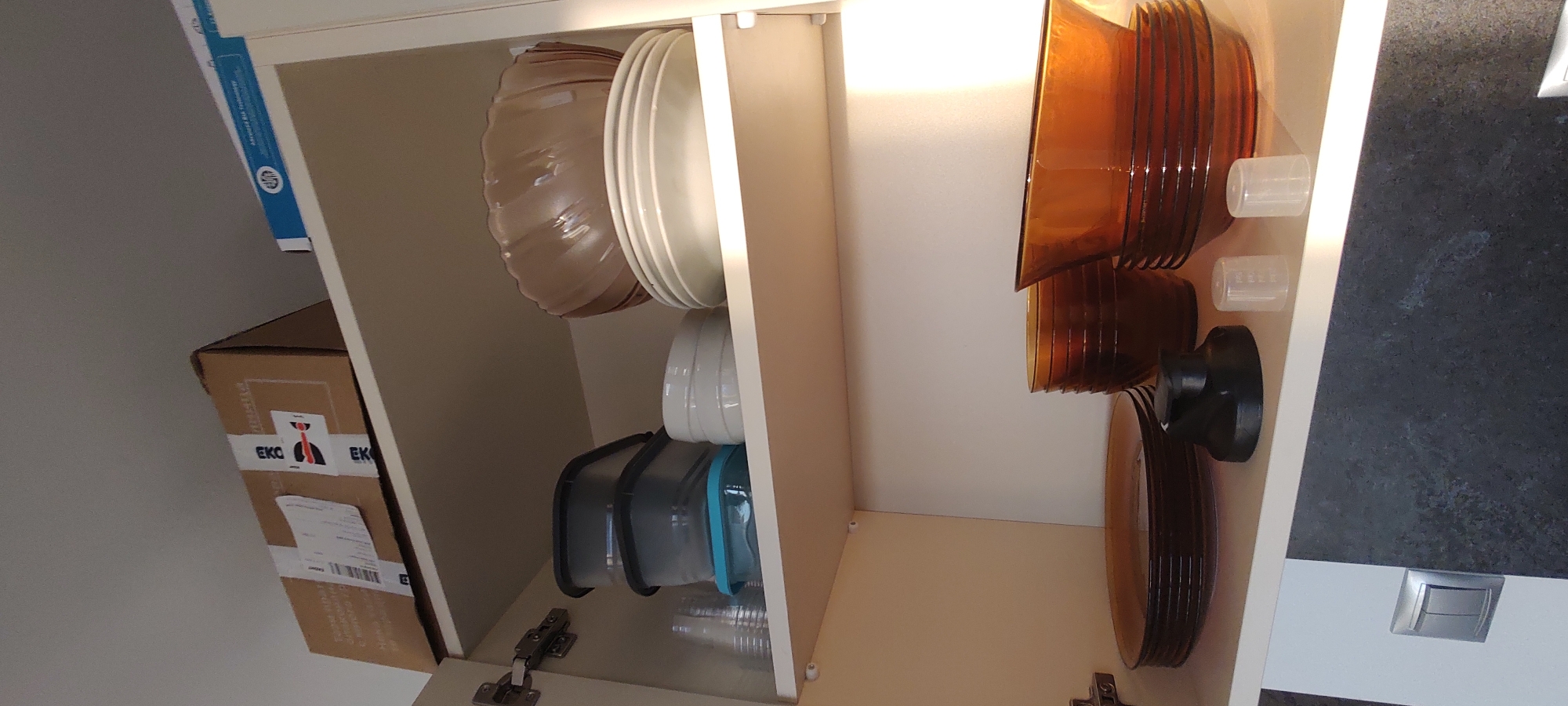 The image shows the interior of a kitchen cabinet in an unknown city. The cabinet is divided into two sections. The left side contains a stack of light-colored plates and bowls, some plastic food storage containers, and a cardboard box labeled "EKC." The right side contains a stack of brown-colored plates and bowls, two small clear plastic measuring cups, and a black object (possibly a kitchen appliance or tool). The cabinets appear to be made of white material. The counter next to the cabinet is black. A light switch is also visible on the right side of the image. The setting is a kitchen, likely in a home or apartment.
