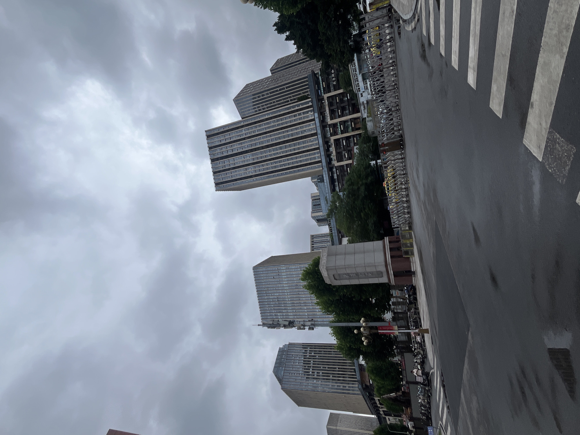 The image shows a street scene in Yuzhong District, China, under an overcast sky. The foreground features a wet asphalt road with a pedestrian crossing. To the right of the crossing, a metal barrier is extended across the road. Behind the barrier, a line of yellow taxis is visible. The middle ground of the image is occupied by several tall, modern buildings with glass facades. A monument or memorial structure stands to the left of the buildings, with trees surrounding it. A street lamp with multiple bulbs is positioned in front of the monument. On the left side of the street, a row of motorcycles is parked. A single person in a light-colored shirt is walking near the motorcycles. The background is dominated by the sky, which is filled with dark, heavy clouds, suggesting inclement weather.