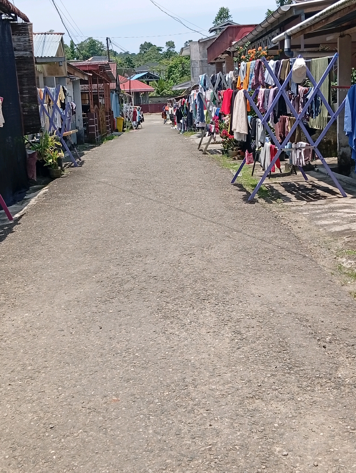 The image depicts a narrow, paved street in the City of Gunungsitoli, Indonesia, lined with homes and clothes drying on racks. On the left side of the street, laundry is hung on purple A-frame drying racks in front of houses with corrugated metal roofs. Potted plants are visible near the houses. On the right side, similar purple drying racks hold a variety of clothing items, and a blue towel hangs from a structure attached to a house. The street itself is made of asphalt and gravel. In the distance, more houses and trees are visible under a bright, partly cloudy sky. A few people are walking in the distance. The scene appears to be a residential area where laundry is being dried outdoors.