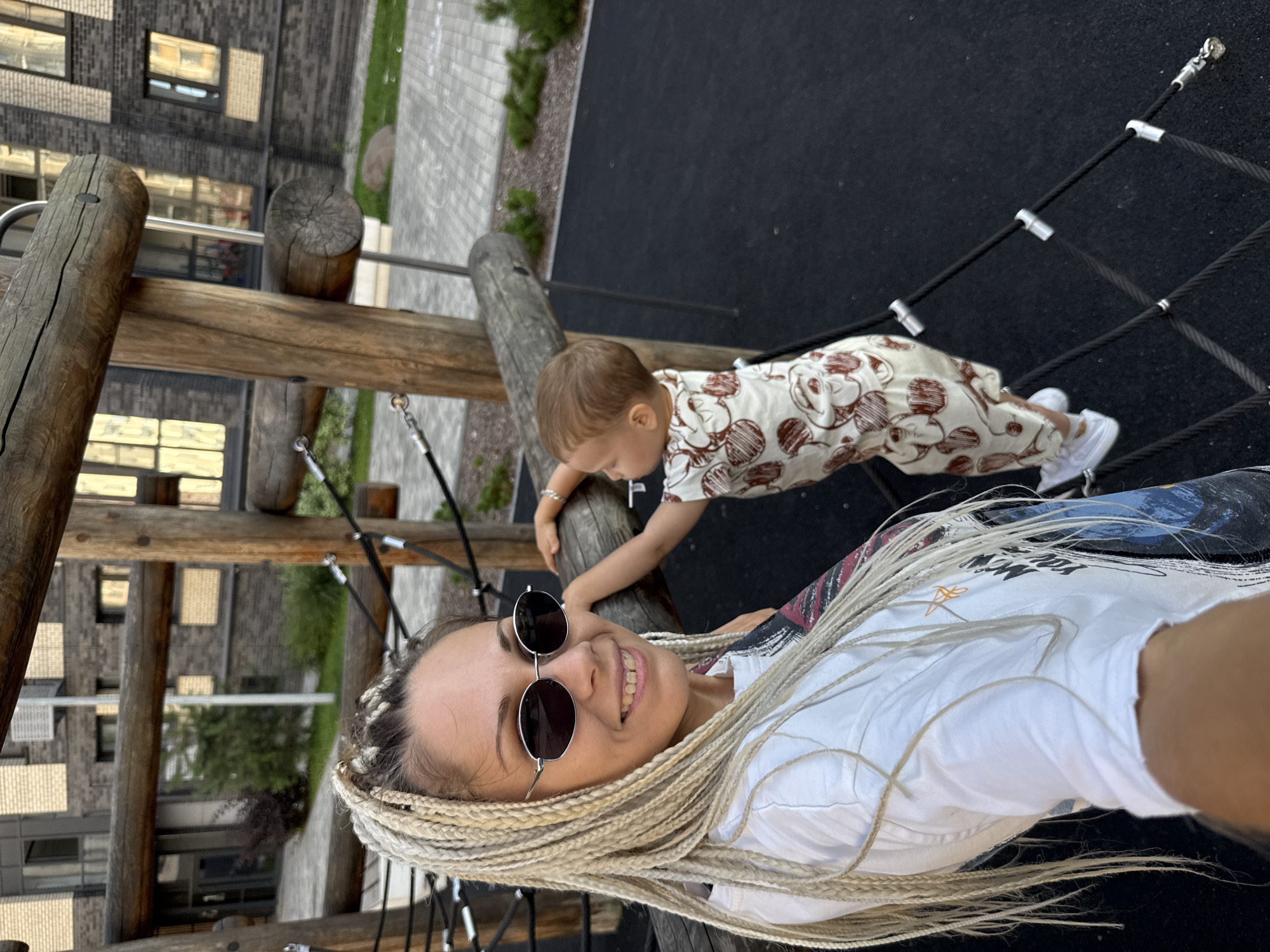 In Rusanovo, Russia, a woman with blonde braided hair and sunglasses smiles for a selfie on a playground. A young child in a patterned outfit is climbing on a wooden structure behind her. The playground has a black rubberized surface and wooden beams with rope netting. Buildings with brick facades are visible in the background.