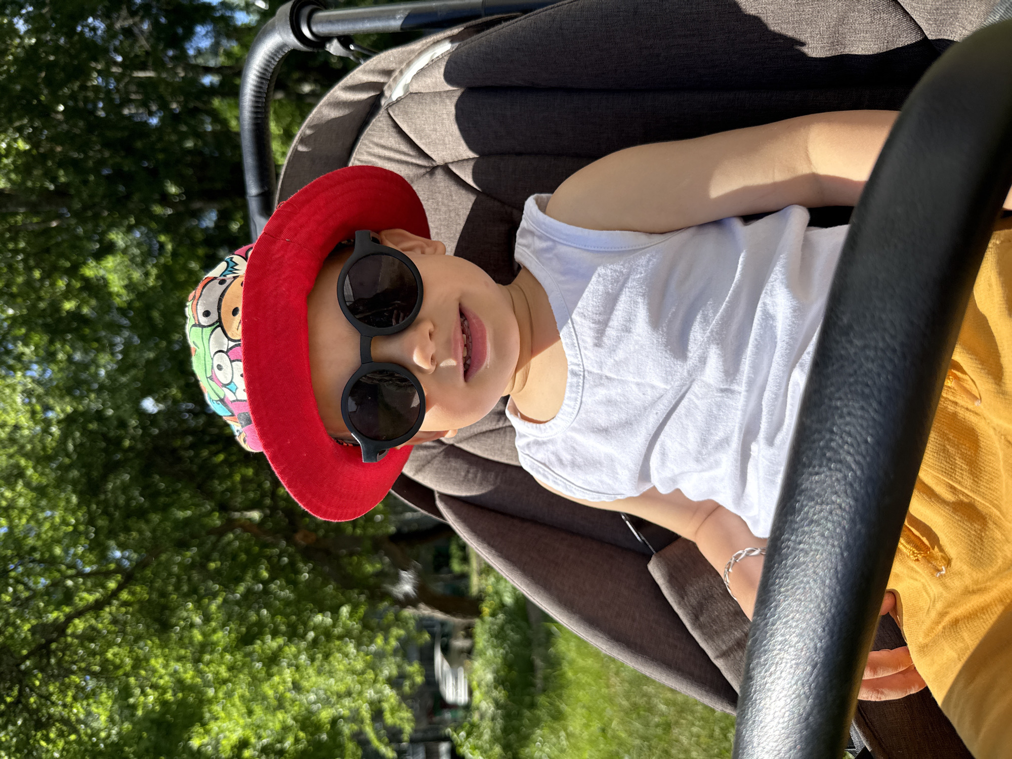 A young child wearing a red hat with cartoon characters, sunglasses, and a white tank top sits in a stroller on a sunny day. The child's legs are crossed, and they are wearing yellow shorts. The stroller is positioned outdoors, with a grassy area and trees visible in the background. The image appears to have been taken in Rusanovo, Russia, as suggested by the context.