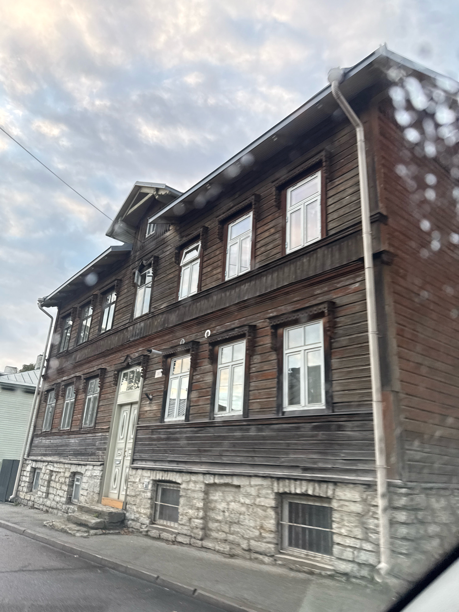 The image depicts a wooden apartment building with stone foundations on a street in Tallinn, Estonia. The building is dark brown, with several windows on each of its visible floors. The windows are white-framed and rectangular, with many reflecting the cloudy sky above. The lower level of the building features a stone foundation with smaller, barred windows. A light-colored door is centrally located on the first floor, accessible by a short flight of stone steps. The street in front of the building is paved, with a concrete curb and sidewalk. The sky is overcast with visible cloud patterns. A power line runs diagonally across the upper left portion of the image. The perspective suggests the photo was taken from a moving vehicle, as evidenced by the slightly blurred foreground and the appearance of water droplets on a windshield in the upper right. No people are visible in the scene. The architectural style of the building is characteristic of older residential structures found in parts of Tallinn.