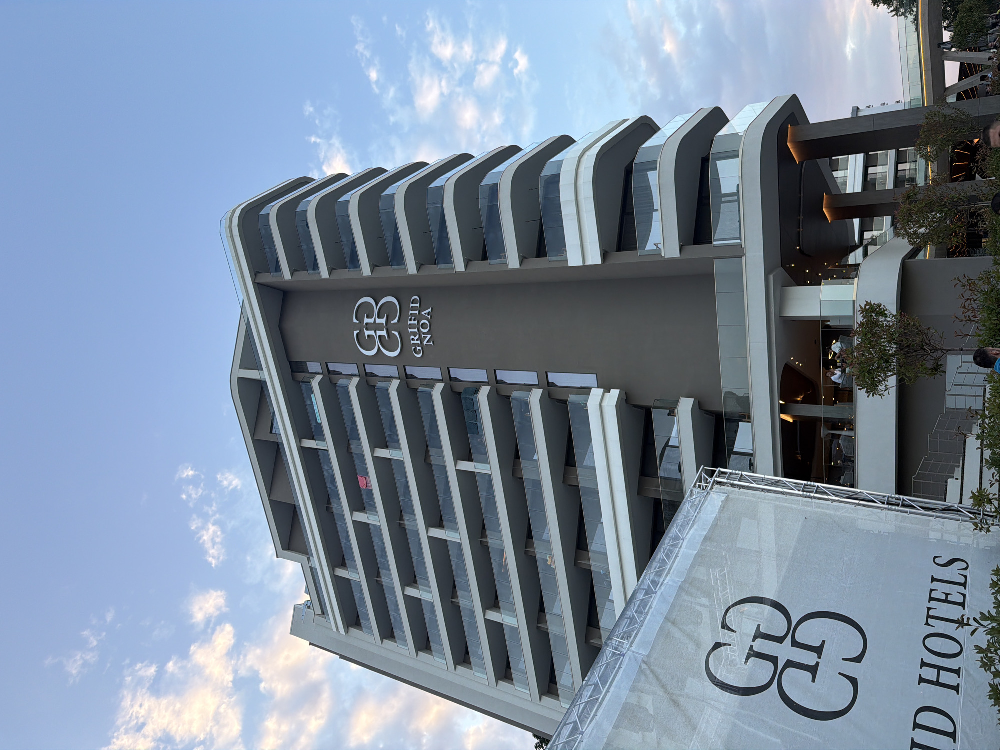 This is a photograph of the Griffid NOA hotel in Varna, Bulgaria. The hotel is a tall, modern building with a dark gray facade and multiple balconies with glass railings. The Griffid logo is prominently displayed on the building's facade. In the foreground, a large white banner with the Griffid logo and the words "ID HOTELS" is visible, supported by a metal frame. The sky is a clear blue with some scattered clouds. The overall impression is one of a contemporary and luxurious hotel.