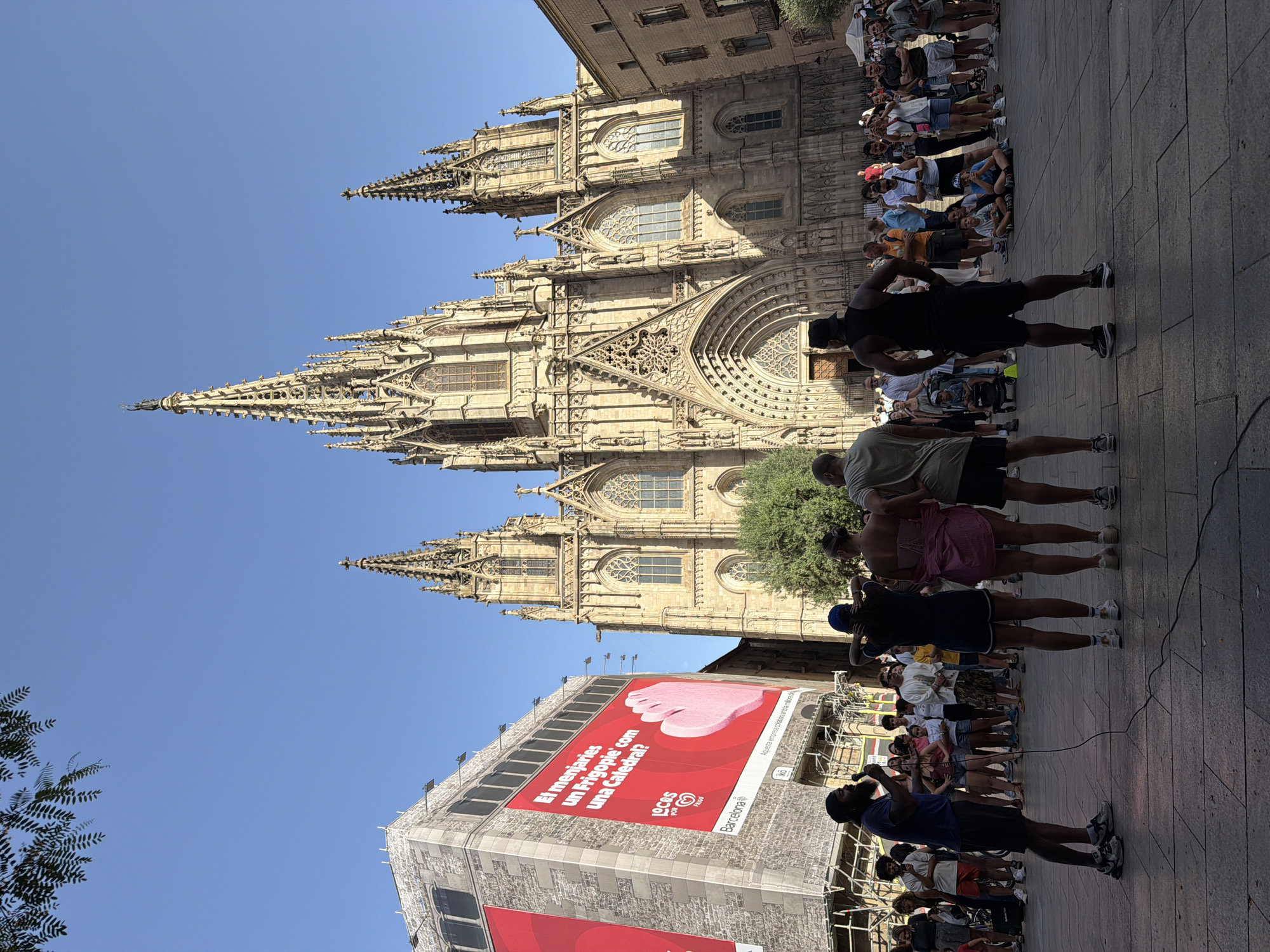 A wide shot captures the exterior of the Barcelona Cathedral on a bright, sunny day, with a clear blue sky above. In the foreground, a diverse crowd gathers on a paved plaza. A man holding a microphone stands on the left, facing a group of three people who are also facing away from the camera. To the left of the cathedral, a large building is covered with a massive advertisement. The architecture of the cathedral is Gothic, with intricate details and soaring spires. The overall scene depicts a public gathering in front of a significant historical landmark in Barcelona, Spain.