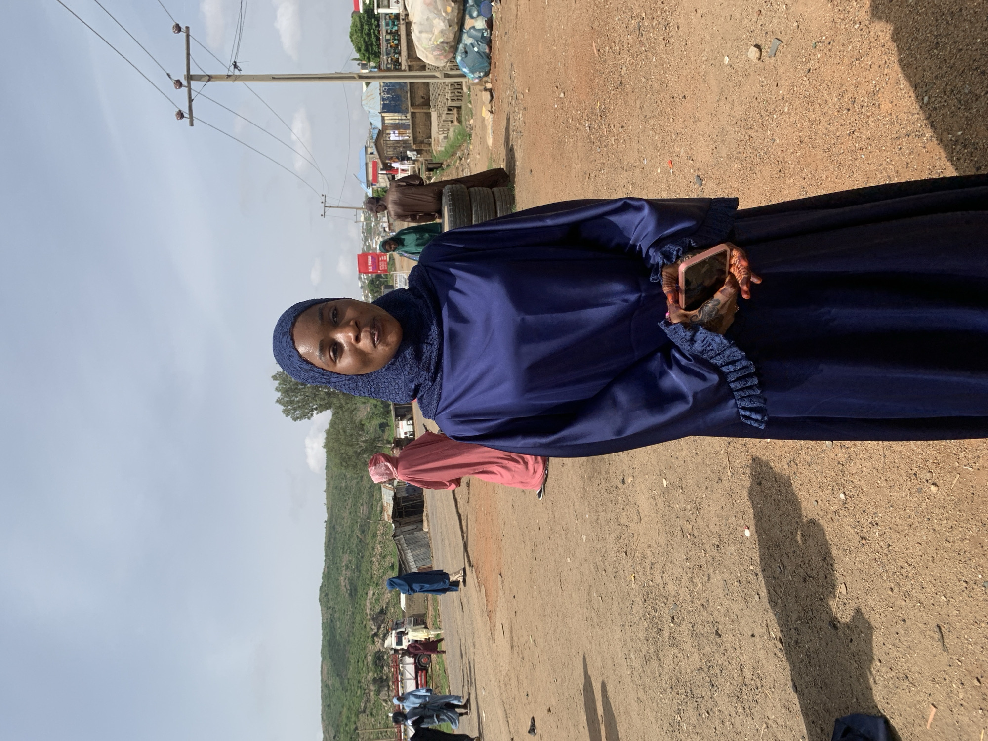 A woman wearing a blue hijab and a long blue dress is standing on a dirt road in Naraguta Hausawa, Nigeria. She is holding a pink cellphone in her left hand. In the background, there are other people walking along the road and some buildings. There is also a green hill on the left side of the road. A row of telephone poles can be seen on the left side of the road. The sky is partially cloudy.